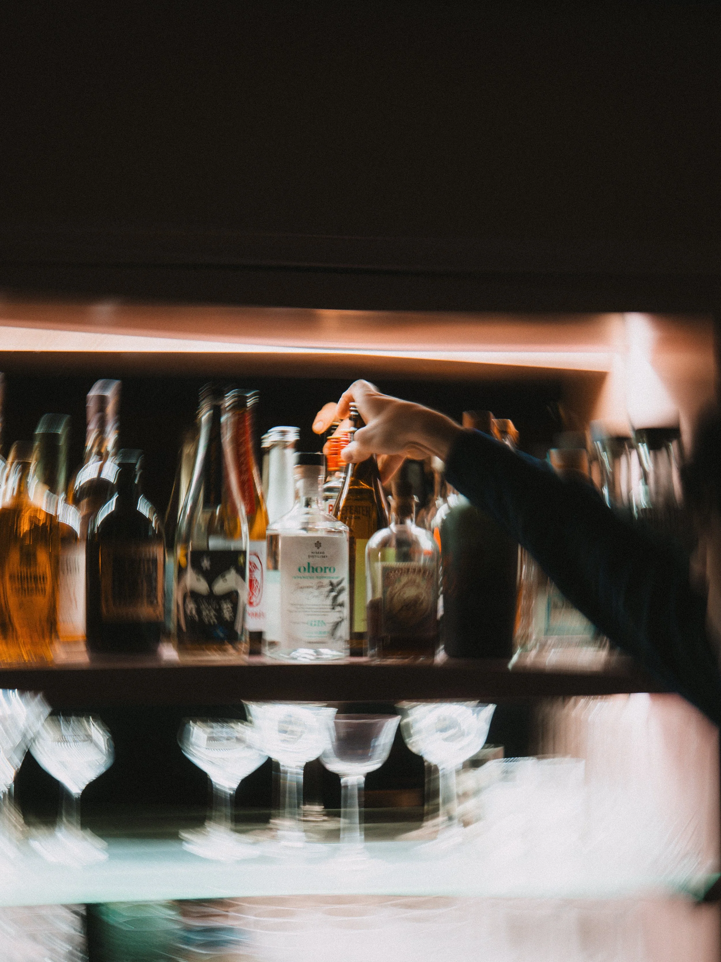 A person reaching for a bottle on a shelf stocked with various liquor bottles in a bar setting.