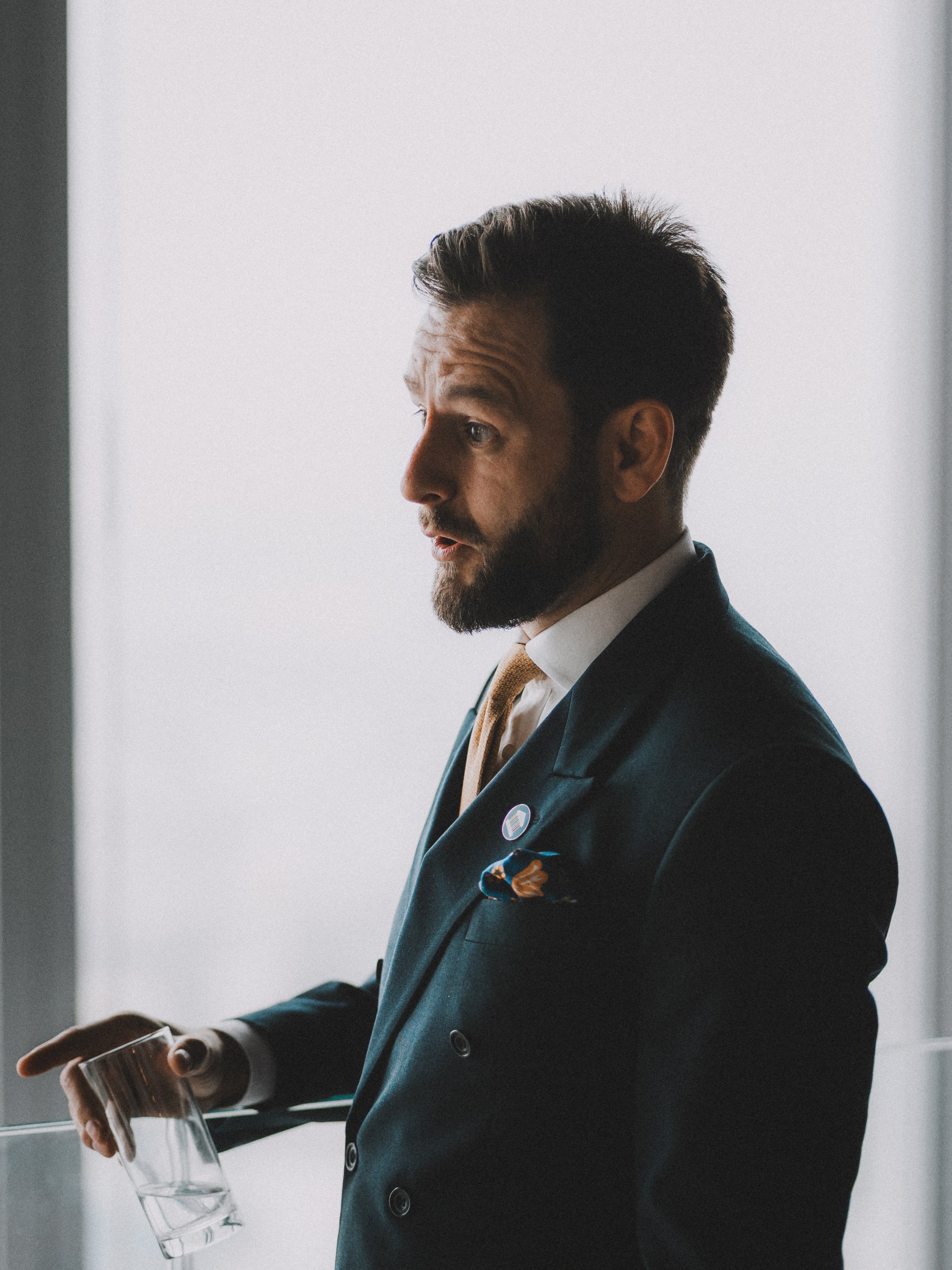 Profile of a man with a beard in a suit holding a glass of water, looking to the side, with a light background.