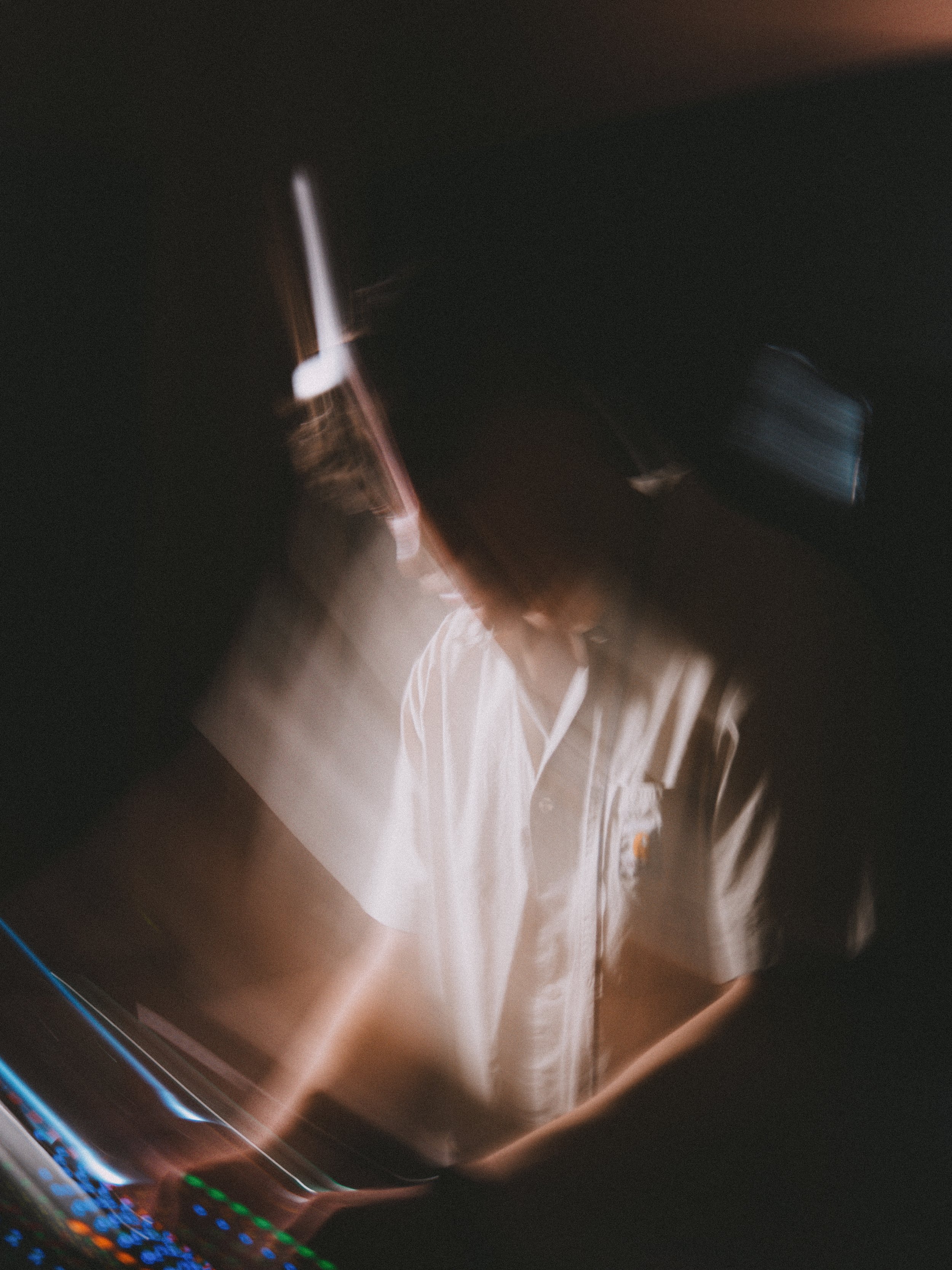 A person with long hair wearing a beige shirt, looking down at a DJ console, illuminated by colorful lights in a dark setting, with motion blur creating an artistic effect.