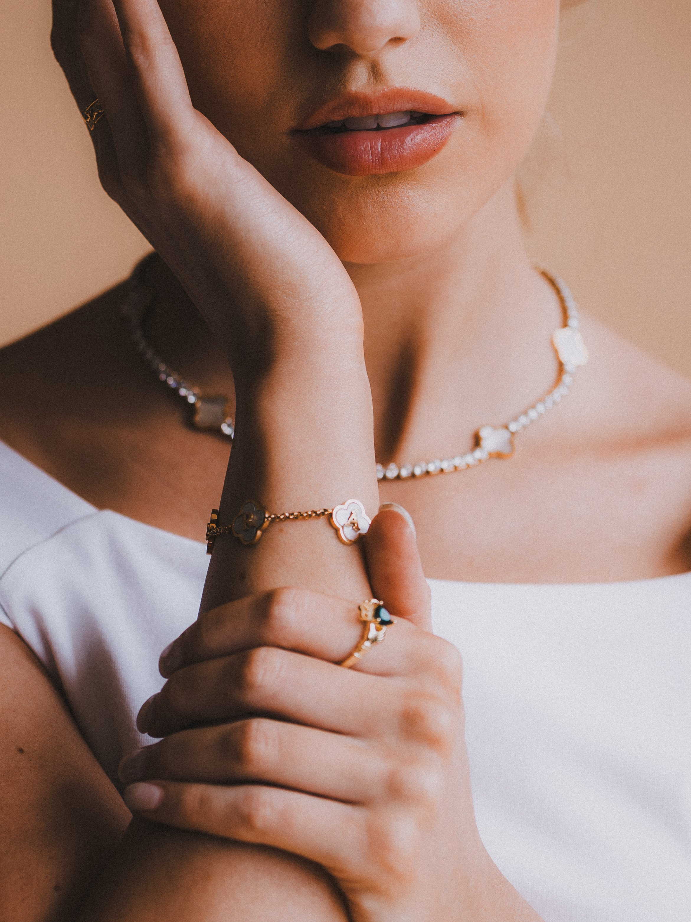 Close-up of a woman wearing jewelry, including a necklace, bracelet, and rings, with her hand gently touching her face and shoulder.