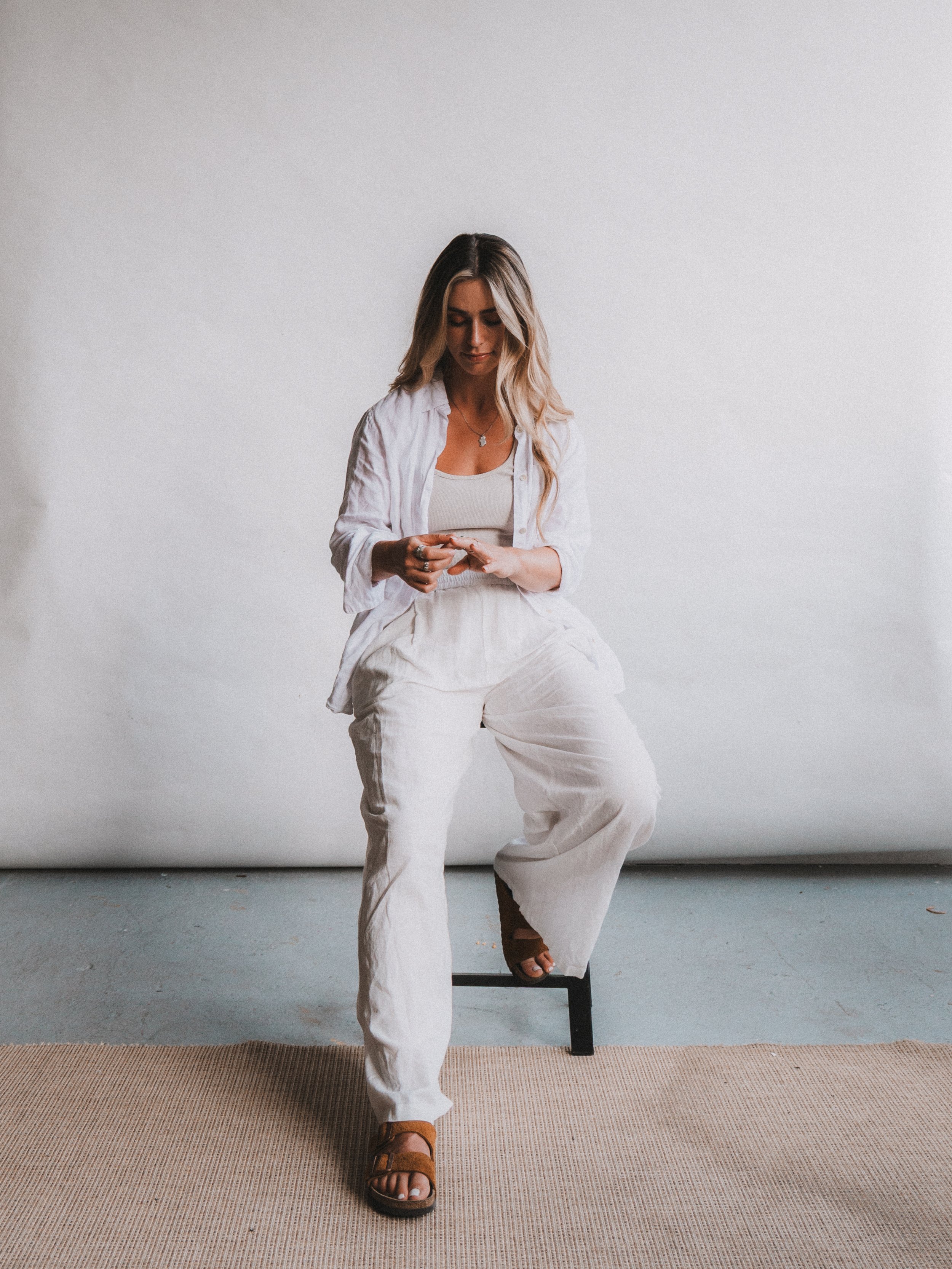 A woman with blonde hair wearing a white shirt and white pants, sitting on a stool against a plain background, looking at her phone.