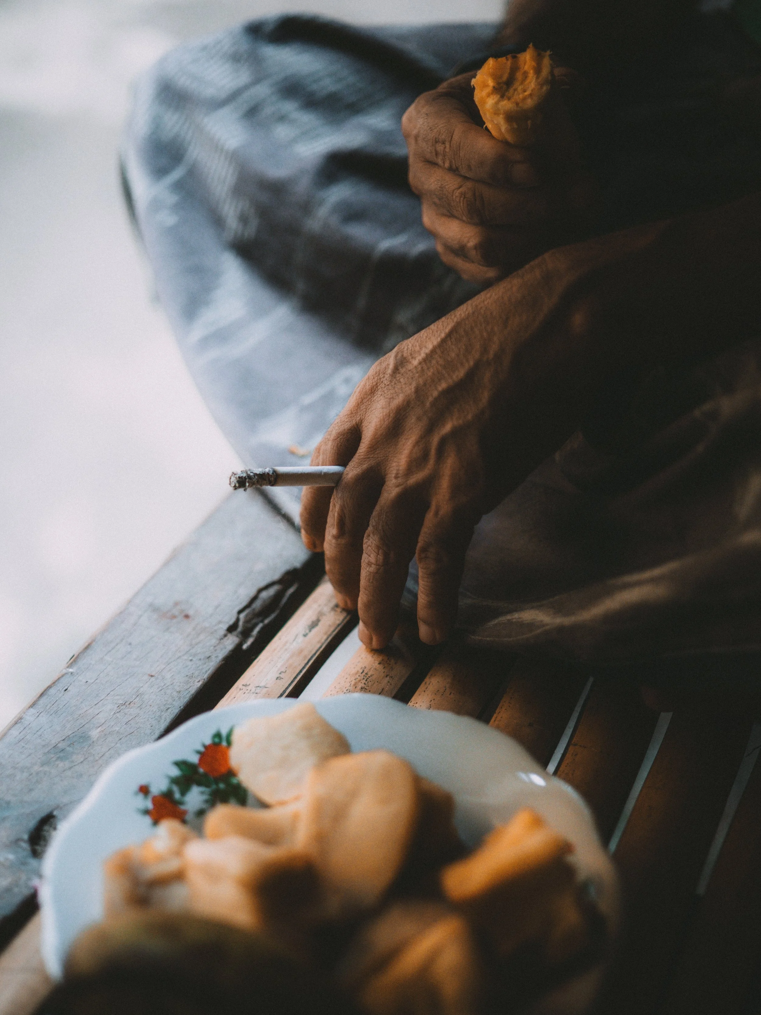 Close-up of a person's hand holding a cigarette and a piece of food, with a plate of fried chips or plantains in the foreground, sitting on a wooden surface.