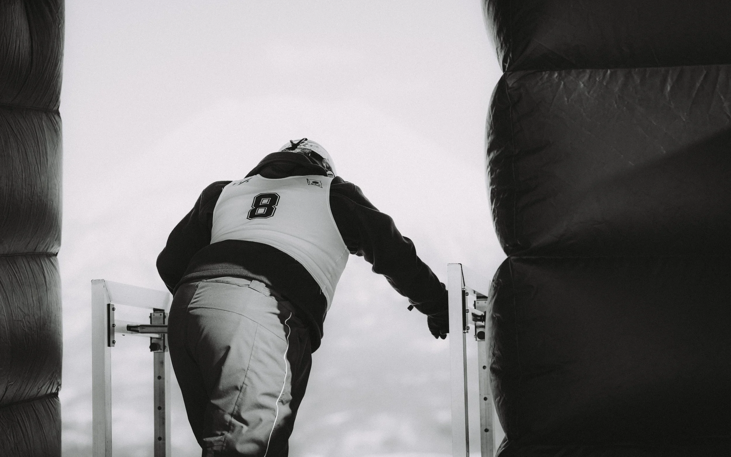 A person wearing a helmet and sports jersey with the number 8 is seen from the back, reaching out towards a barrier at a sports event, possibly a race or competition, in a black-and-white photograph.