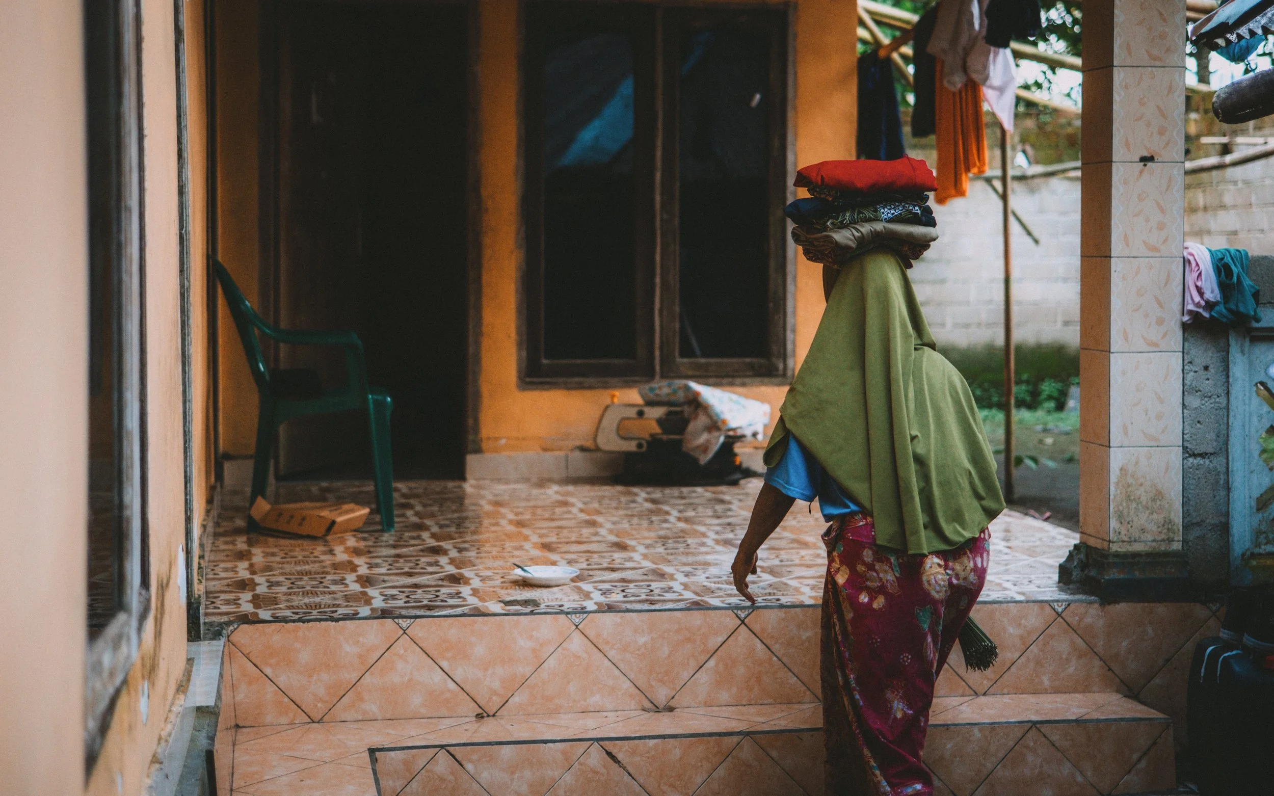 A woman with a green shawl on her head and floral skirt, walking on tiled steps outside a yellow building with a window. Clothes are hanging to dry and a green plastic chair is visible nearby.