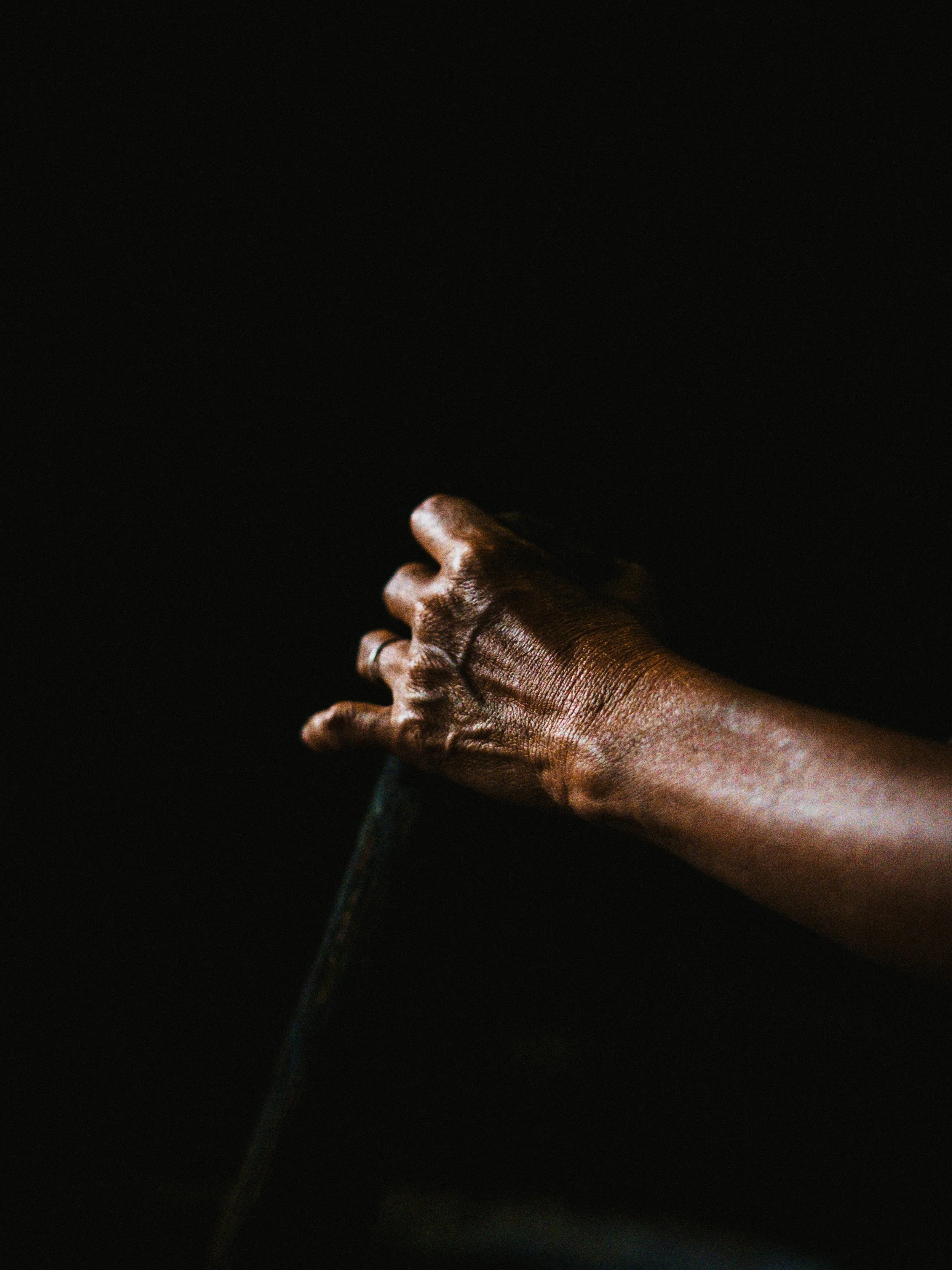 A close-up of a dark-skinned hand gripping a dark object against a black background.