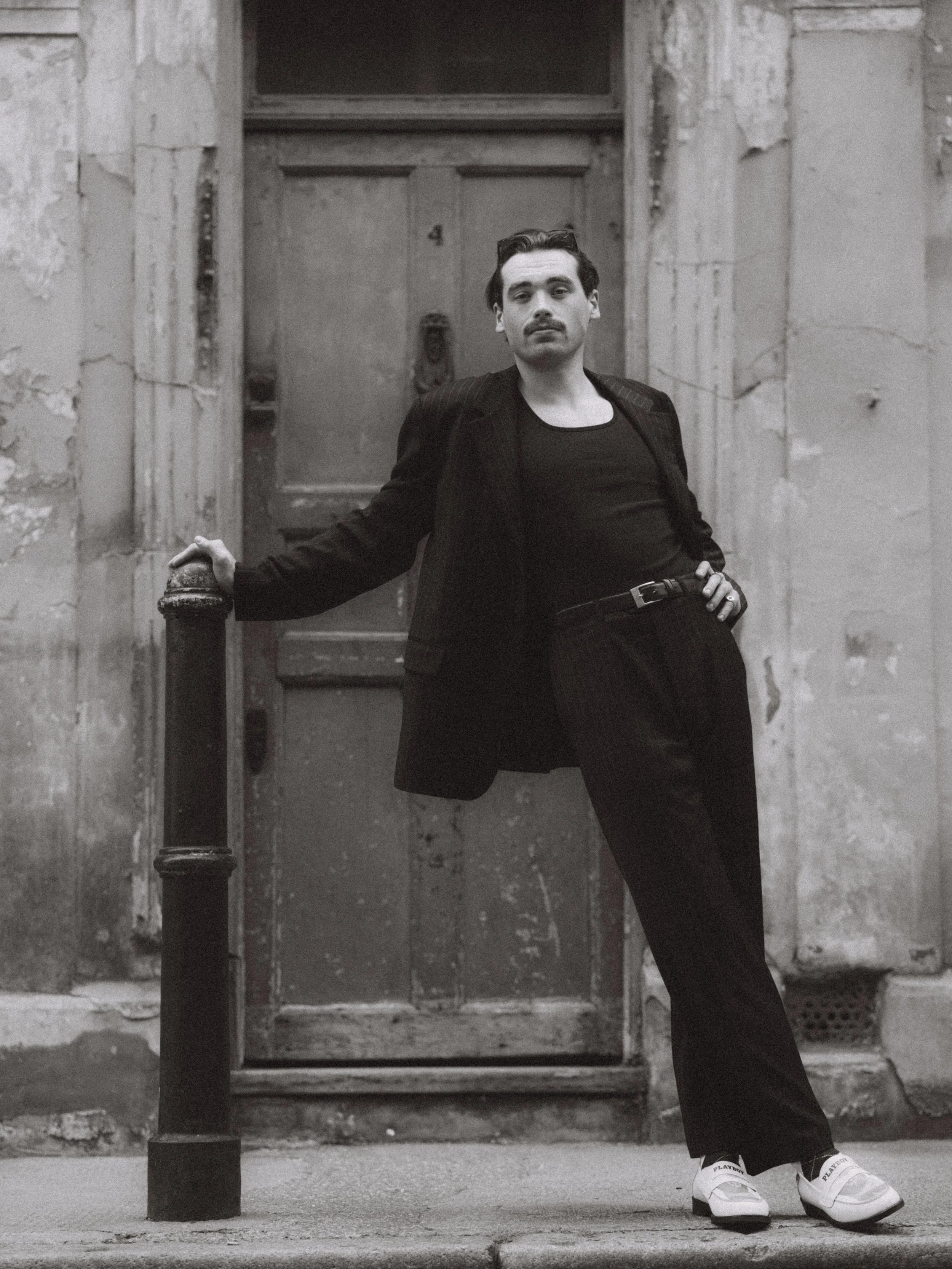 Black and white photo of a man with mustache and dark hair leaning against a post on a city street, wearing a suit jacket, black shirt, and casual sneakers, standing in front of a worn door with peeling paint.