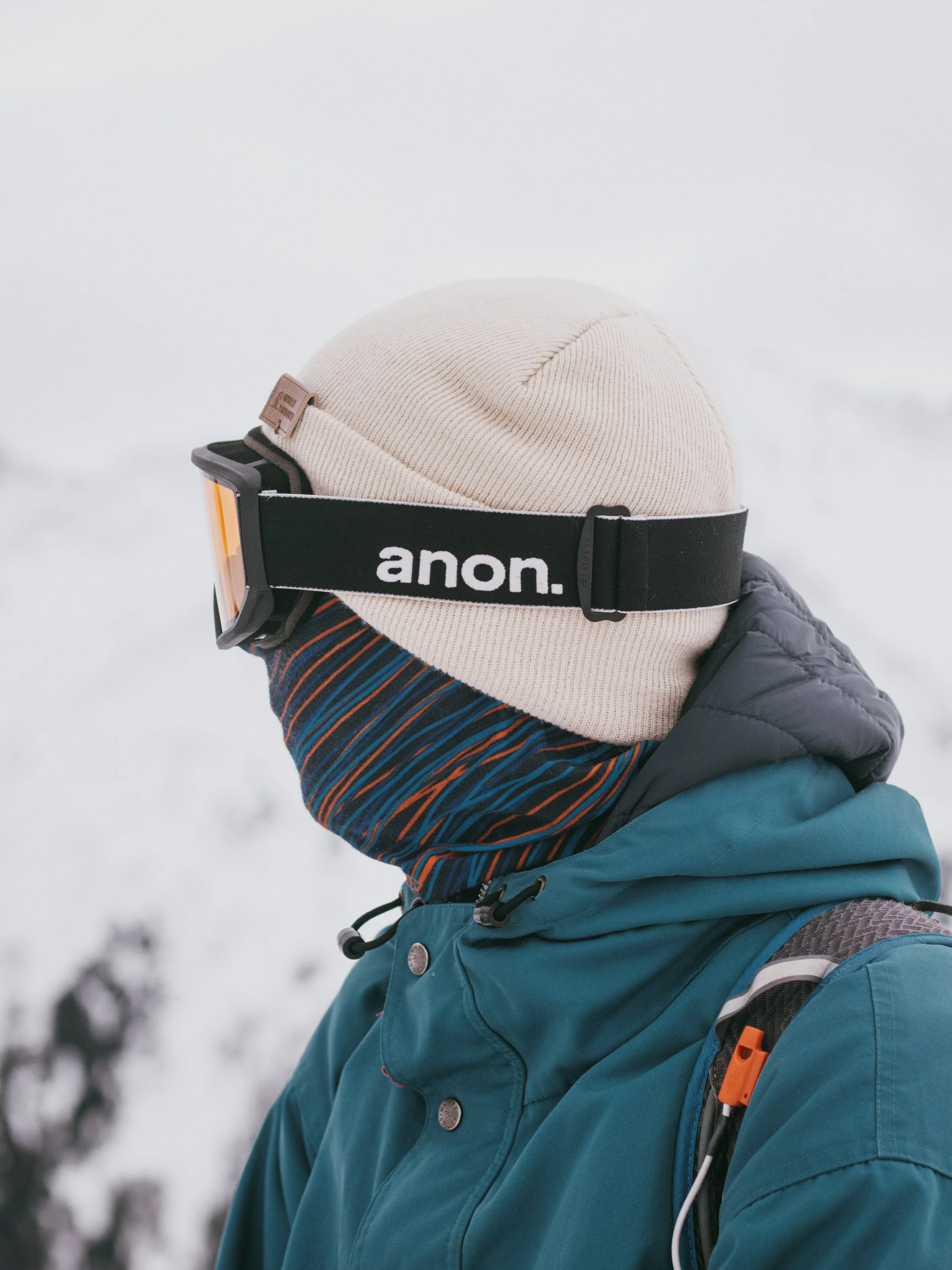 Person dressed in winter gear, wearing a beige beanie, black ski goggles, a multicolored neck gaiter, a teal winter jacket, and a backpack, in a snowy outdoor setting.