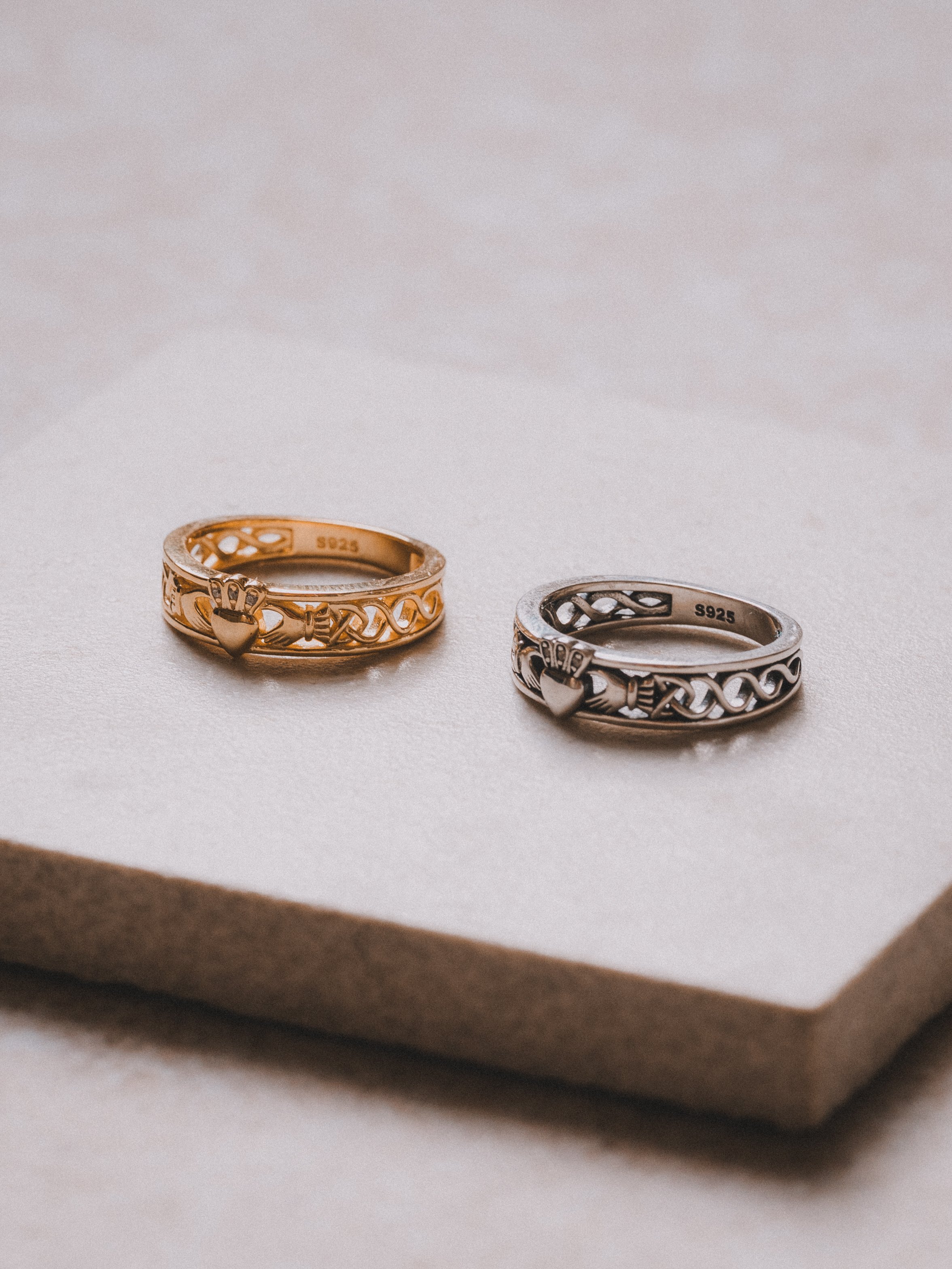 Two decorative rings, one gold and one silver, with heart and crown designs, placed on a textured cream-colored surface.