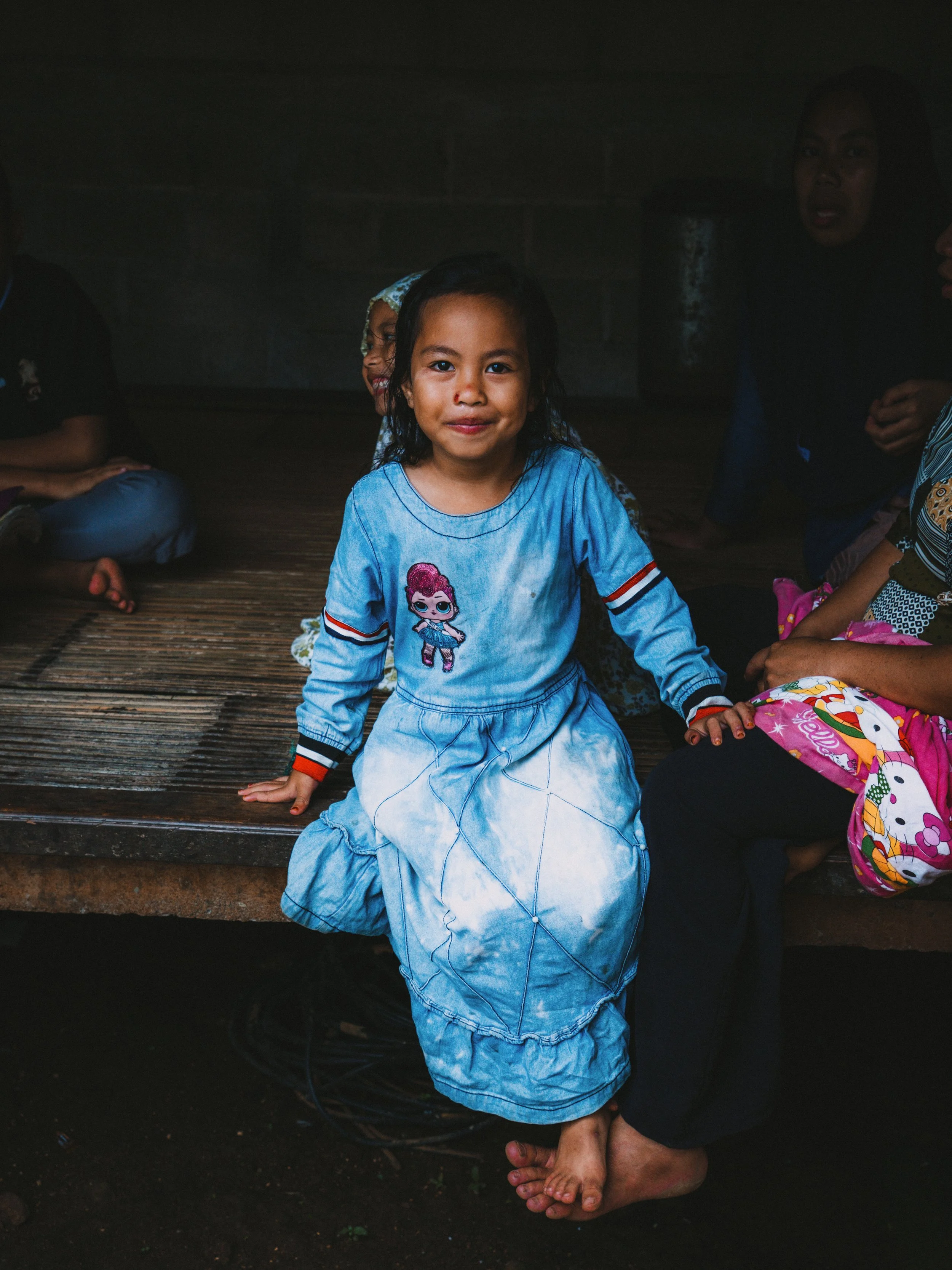 A young girl with dark hair and a blue dress sitting on a wooden platform, surrounded by other children and women in a dimly lit indoor setting.