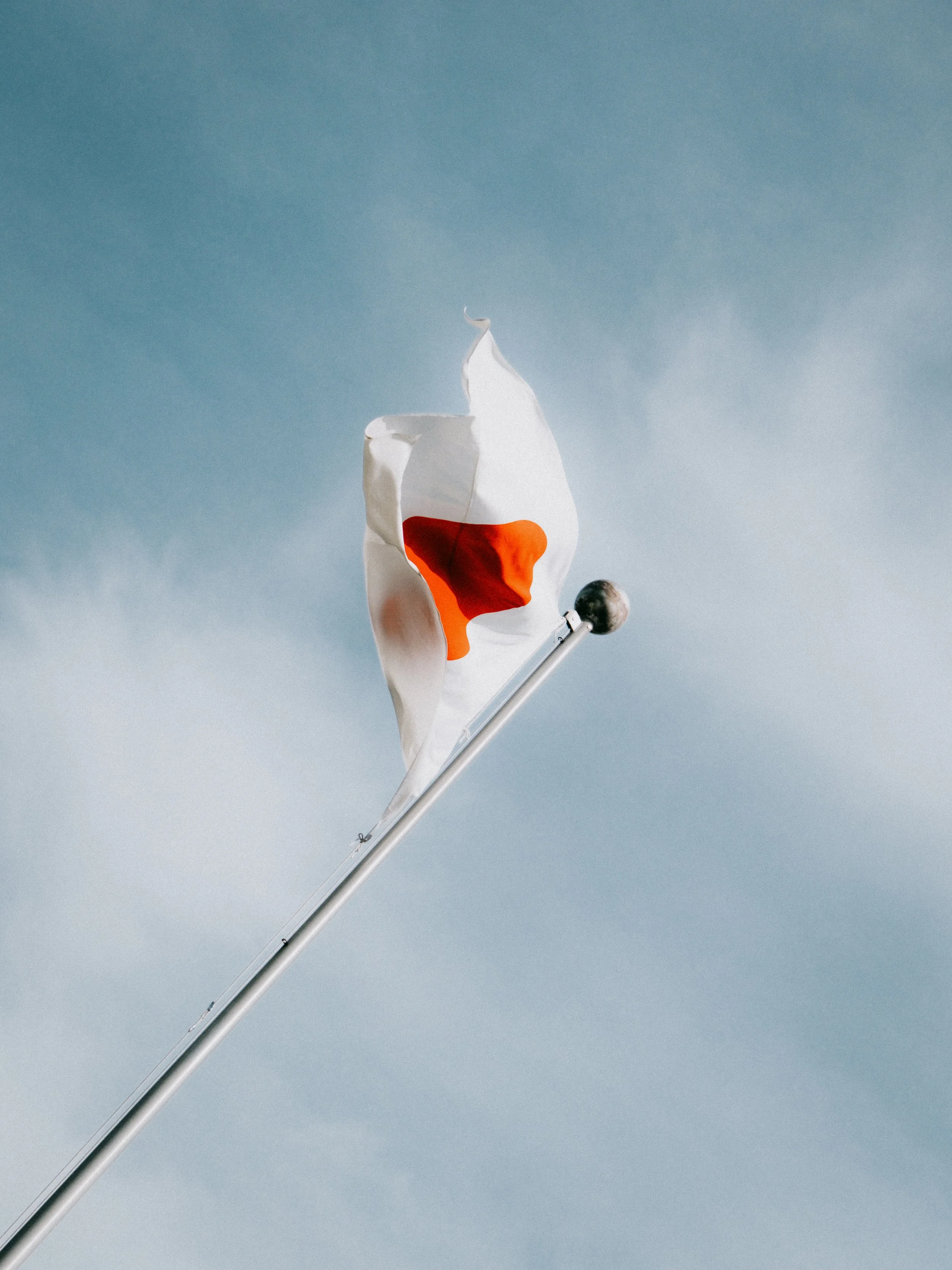 The Japan national flag, with a red circle on a white background, flying on a flagpole against a cloudy sky.