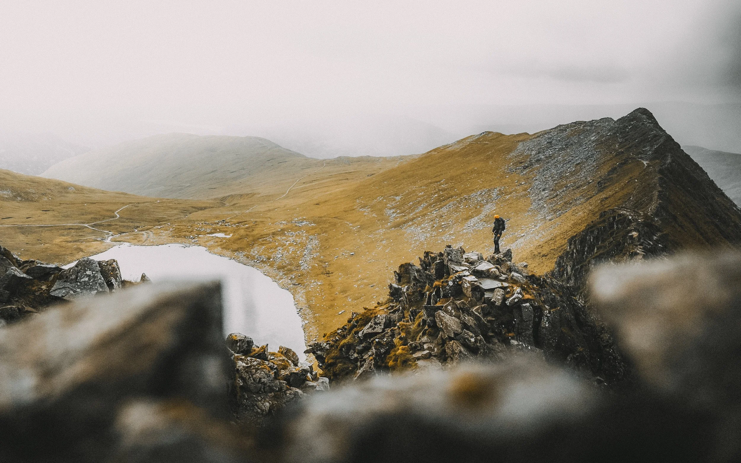 Hiker standing on rocky ridge overlooking a mountain landscape with a lake and rolling hills, some with pathways, under overcast sky.
