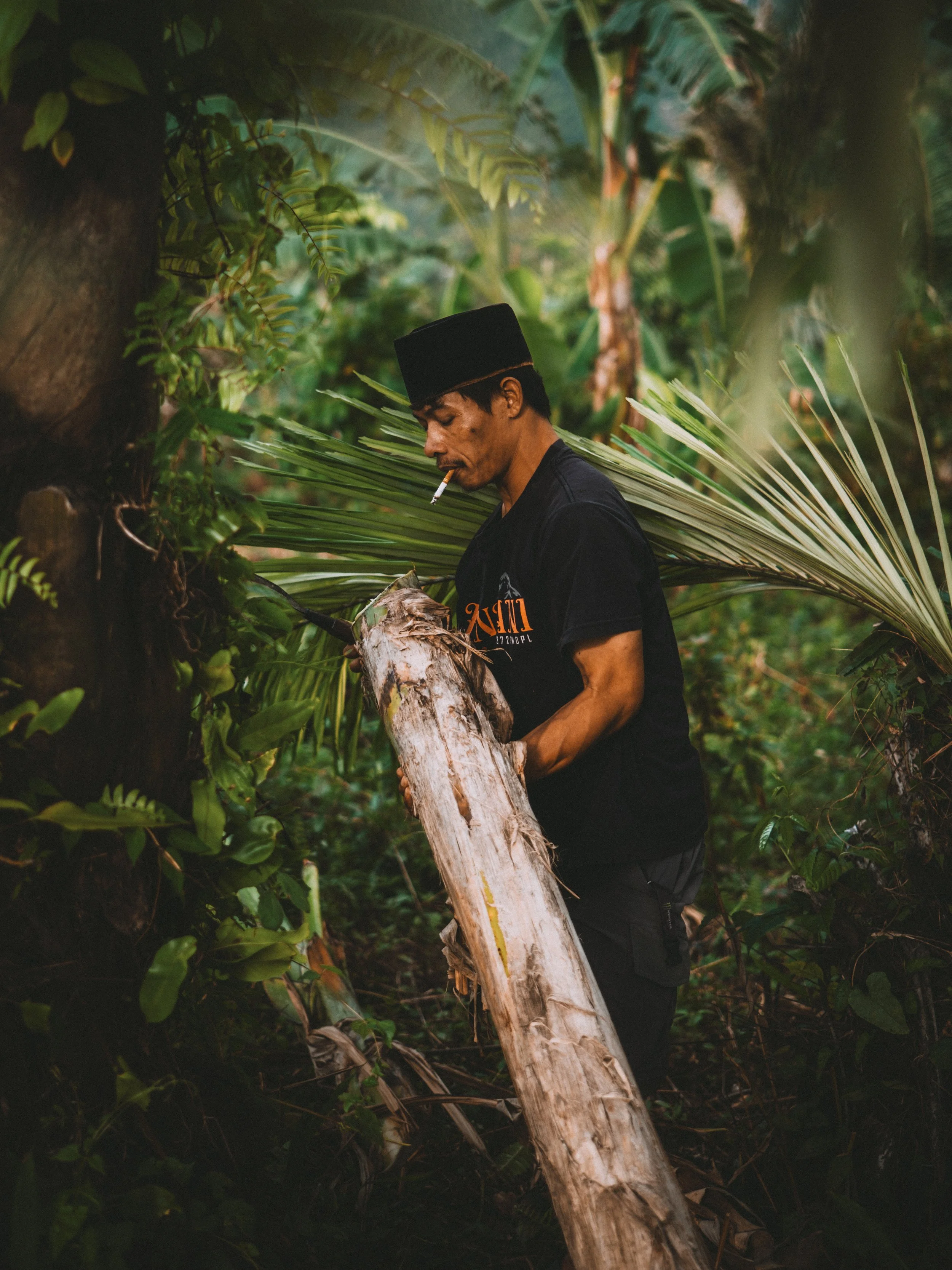 A man in a black shirt and traditional hat is standing in a lush, green jungle, smoking a cigarette, and holding a large, fallen tree trunk.