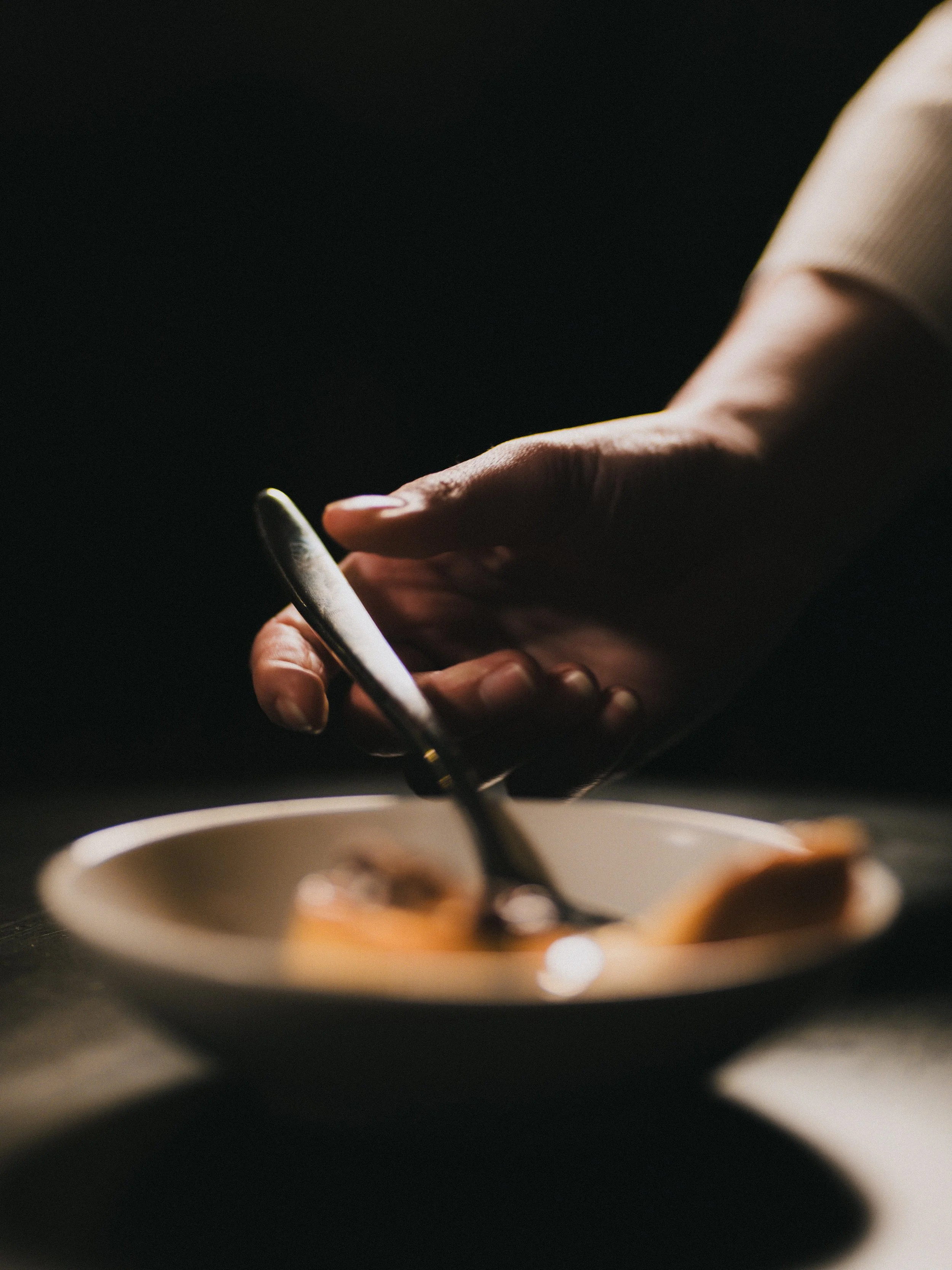 A hand holding a smartphone above a plate with food, possibly dessert, with a spoon resting on it, in a low-light setting.