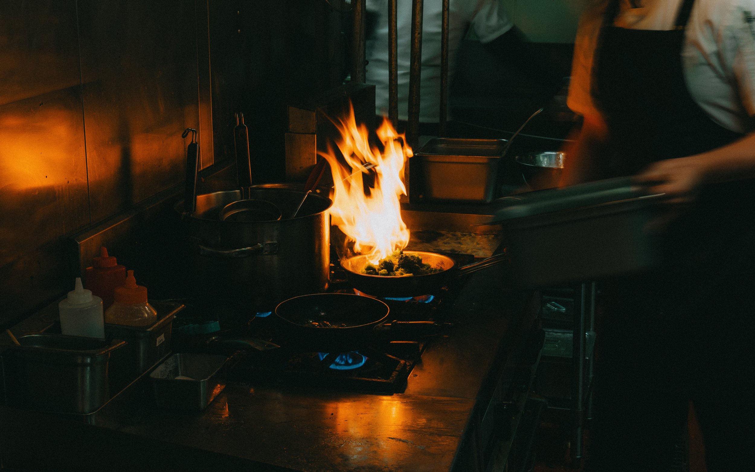 A chef cooking on a stove with a visible flame, surrounded by kitchen utensils and containers.