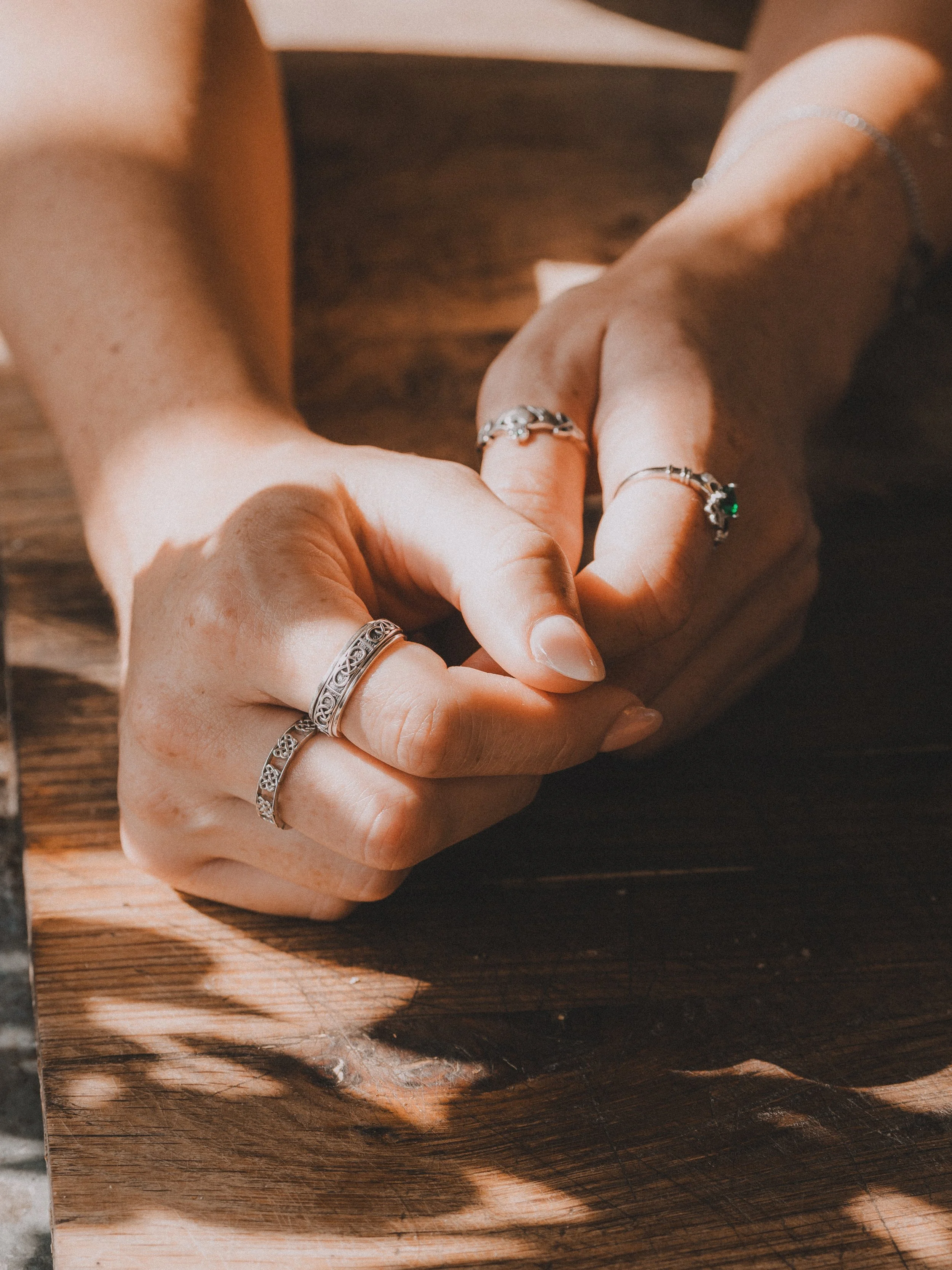 Close-up of a person's hands resting on a wooden table, adorned with multiple silver rings and a silver bracelet, with sunlight casting shadows.