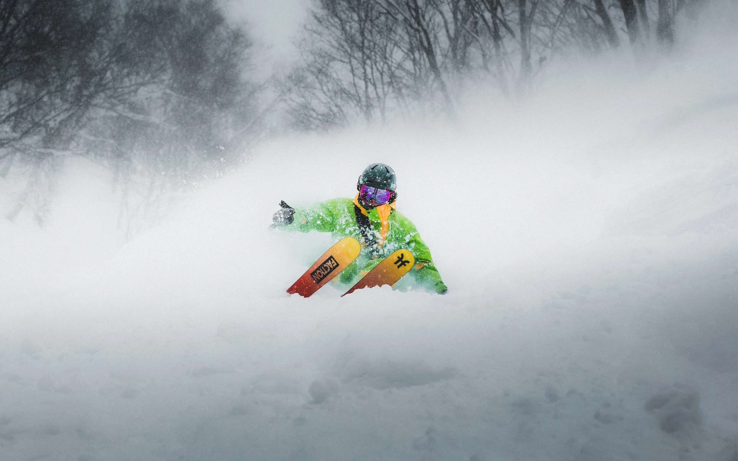 A person wearing a black helmet, purple goggles, and a bright green jacket snowboarding in deep snow with snow-covered trees and a cloudy sky in the background.