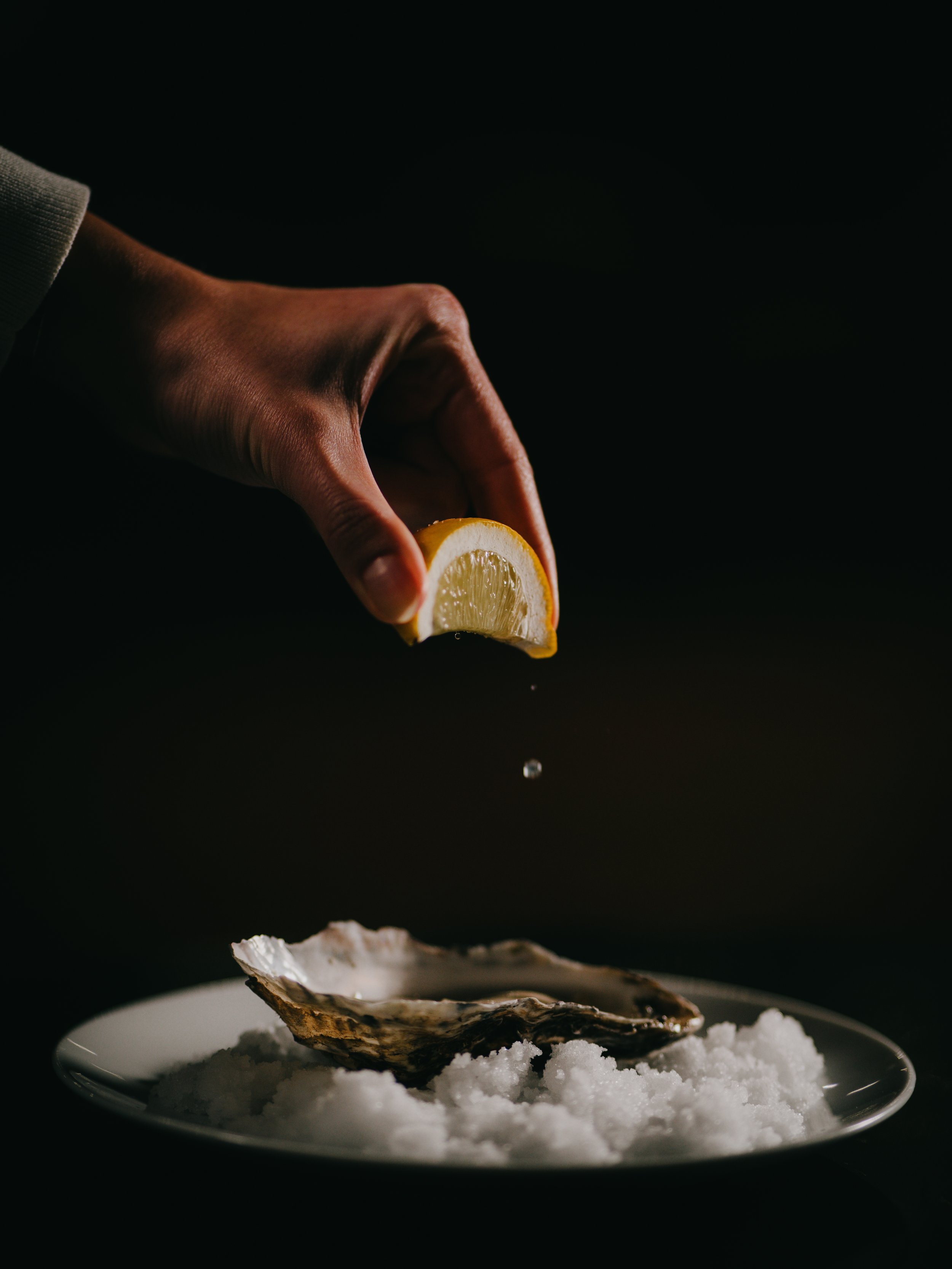 A hand squeezing lemon over a plate of oysters on a bed of ice against a dark background.