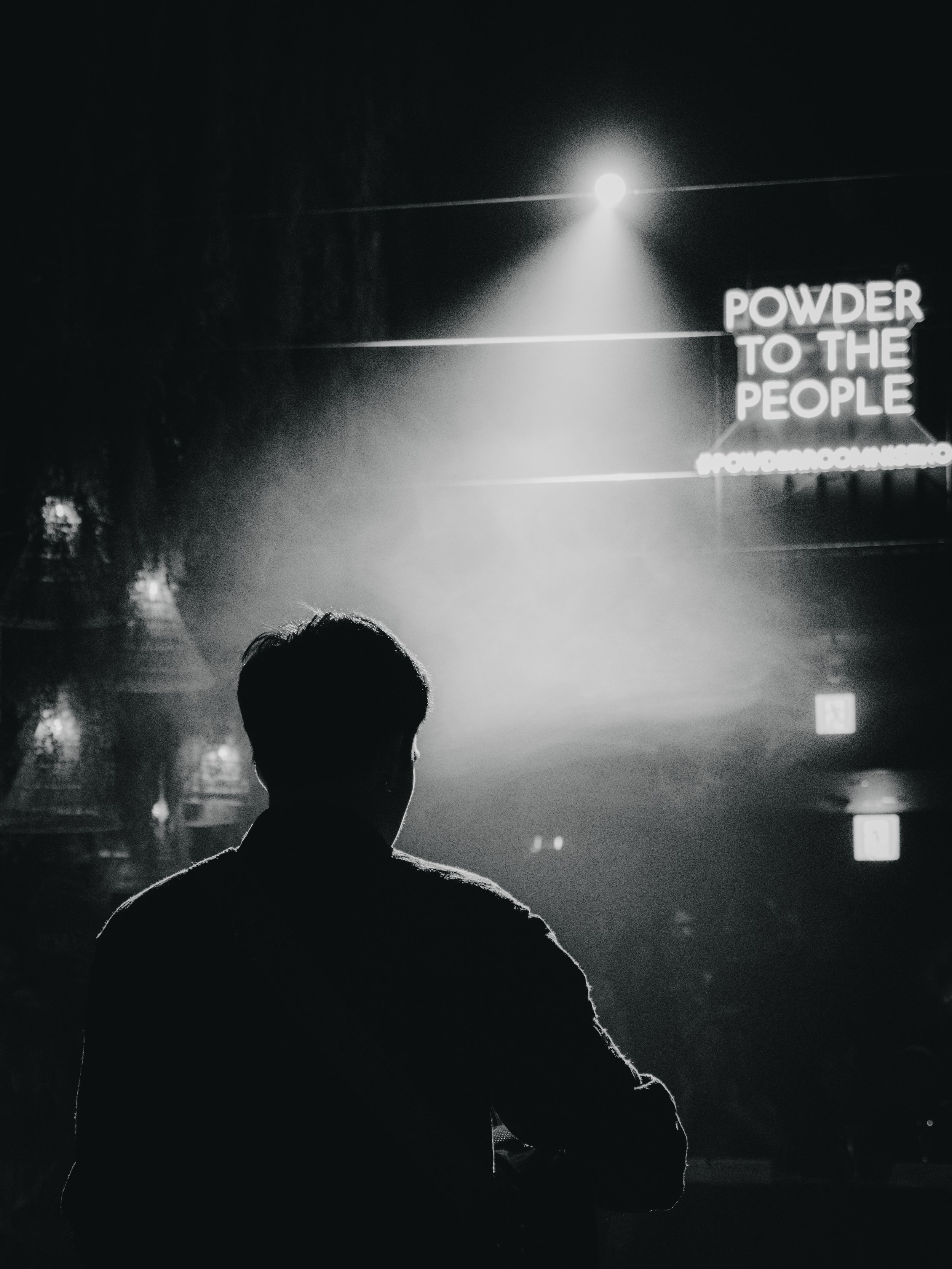 A silhouette of a person standing in front of a stage with a bright spotlight, and a neon sign that reads 'POWDER TO THE PEOPLE' in a dark, indoor venue.