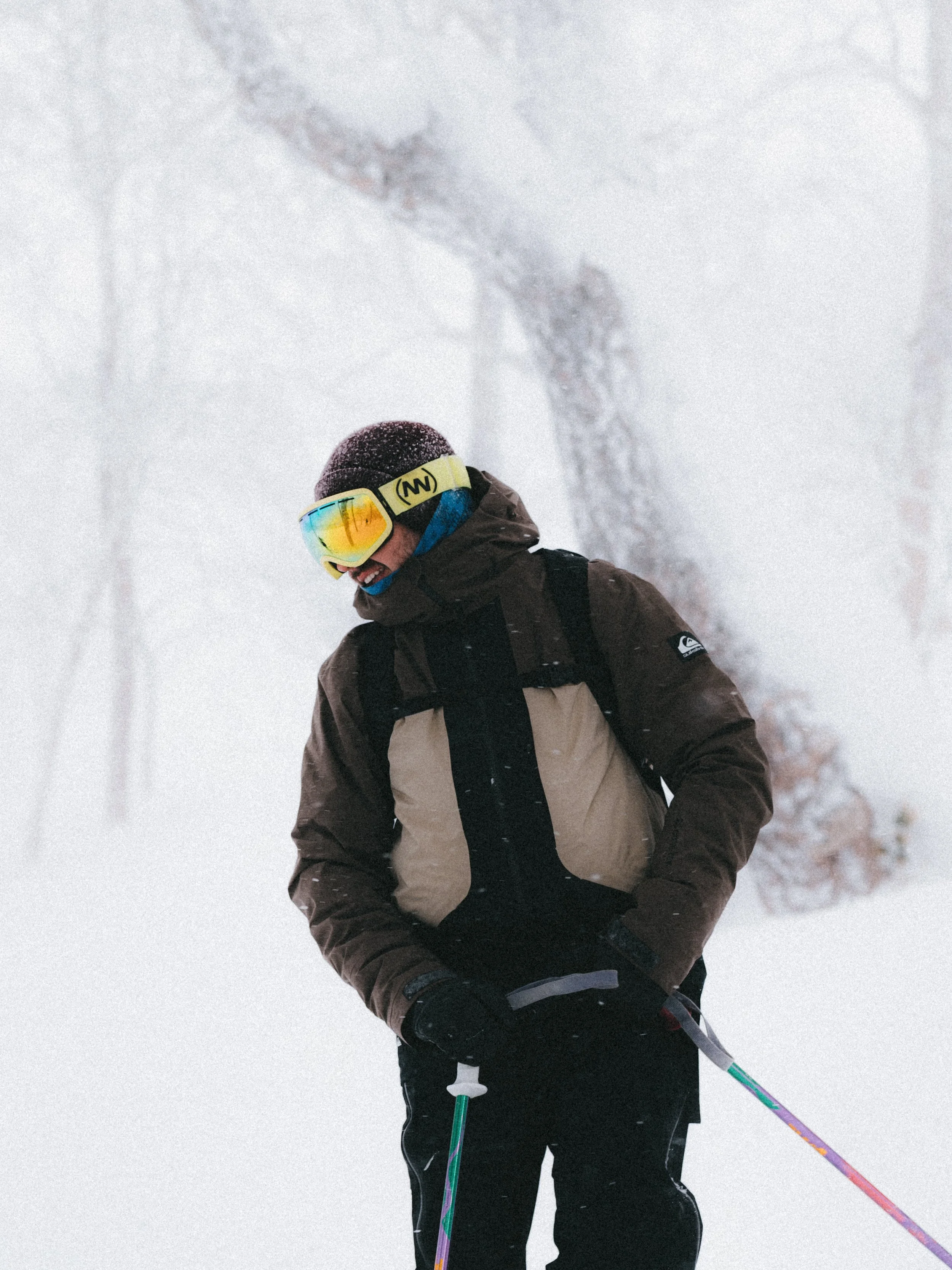 A person in winter gear, including a brown and beige jacket, black pants, gloves, and colorful goggles, standing in a snowy forest with snow-covered trees in the background.