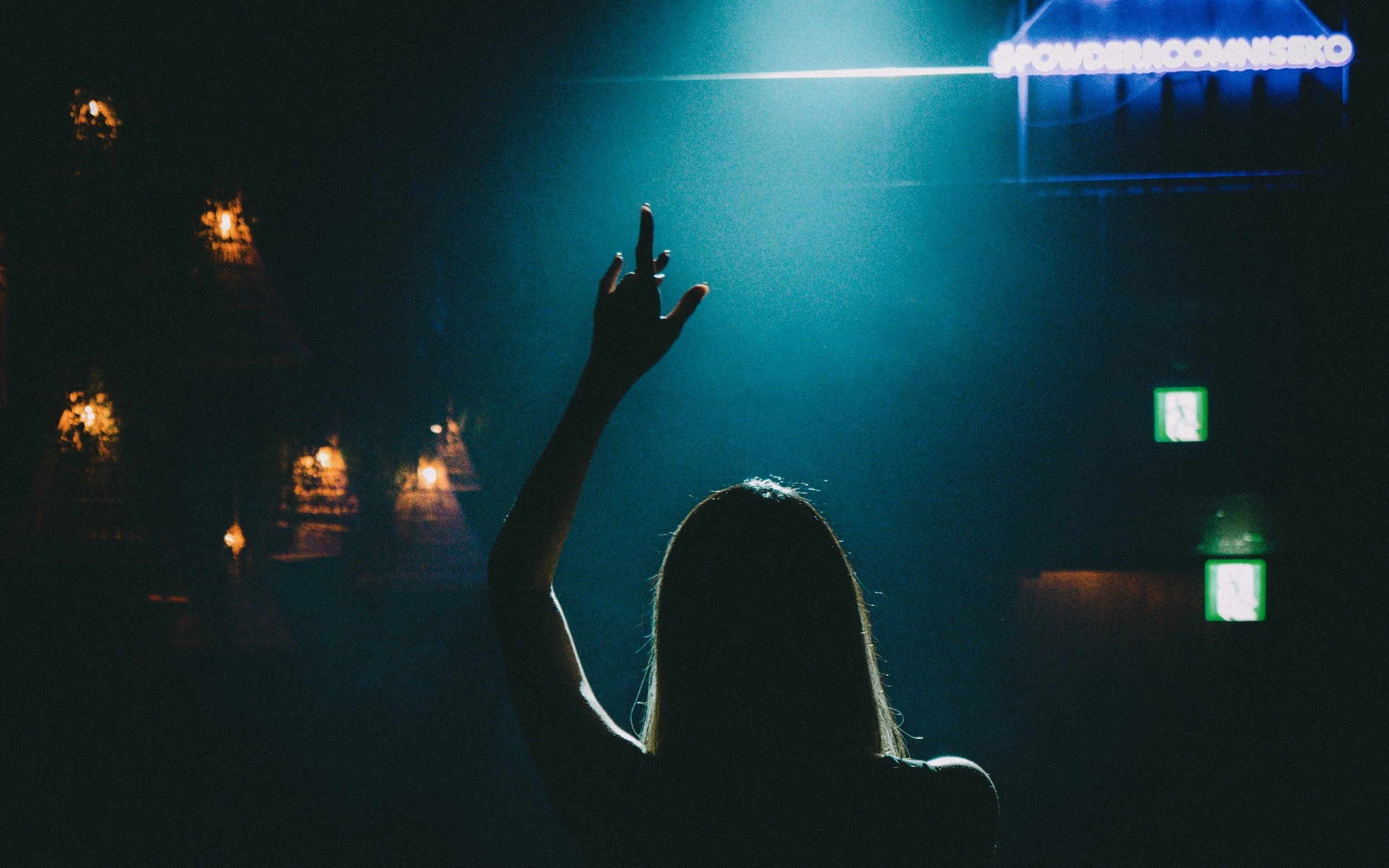 A woman raising her hand in a dark room with stage lights and exit signs visible in the background.