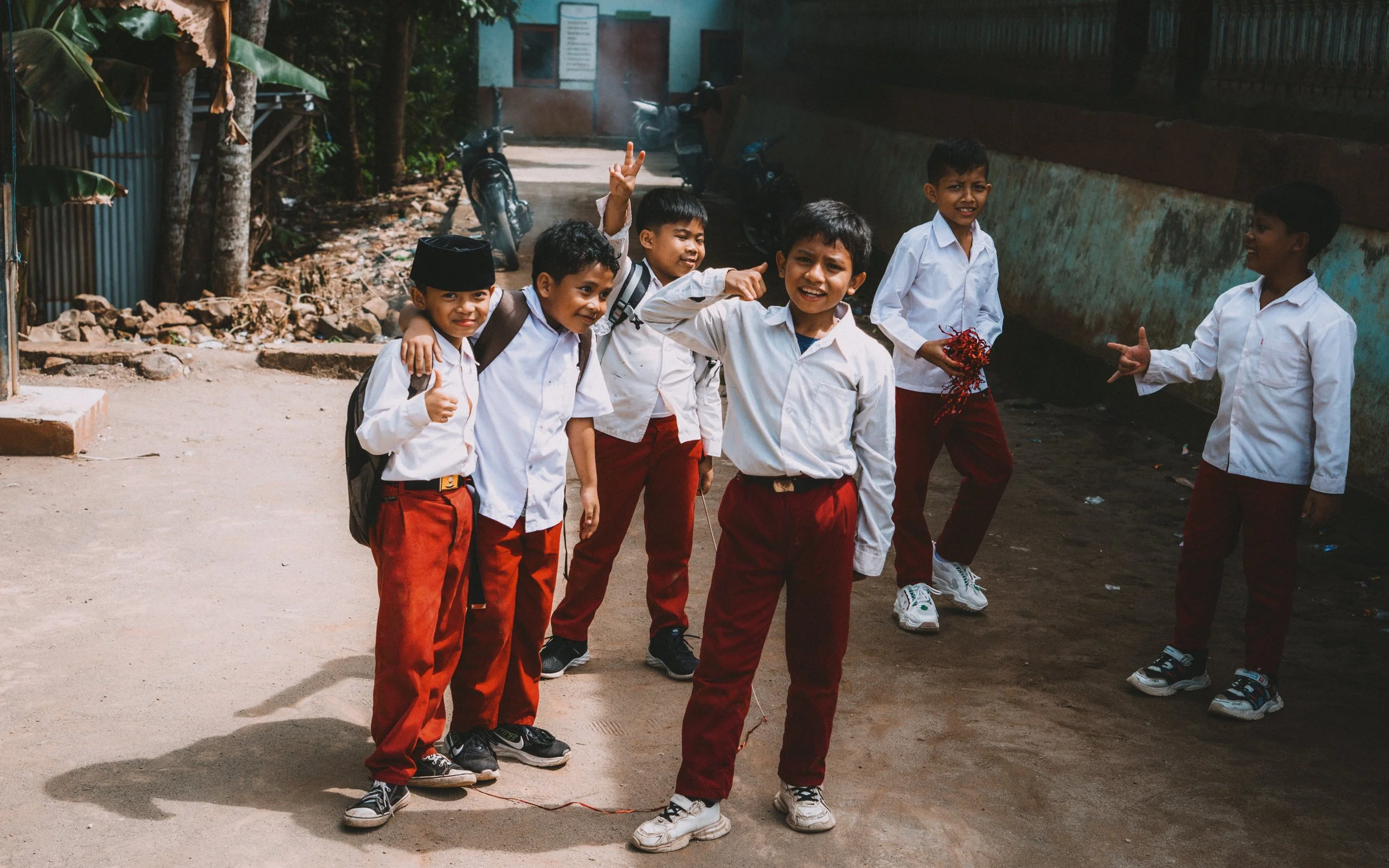 Group of seven schoolboys in white shirts and red pants standing outdoors, smiling and posing for the camera, some making gestures, with trees and a motorcycle in the background.