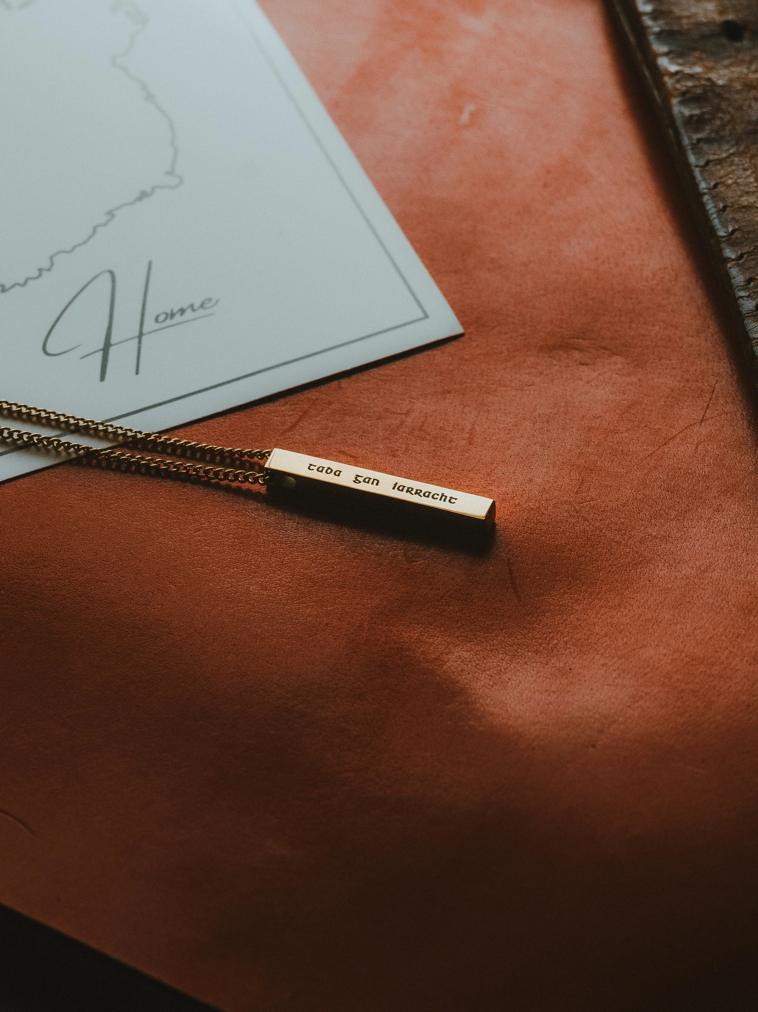 A necklace with a rectangular pendant inscribed with the text 'tada san larrache' resting on a reddish leather surface, next to a piece of paper with the word 'Home' written in cursive.