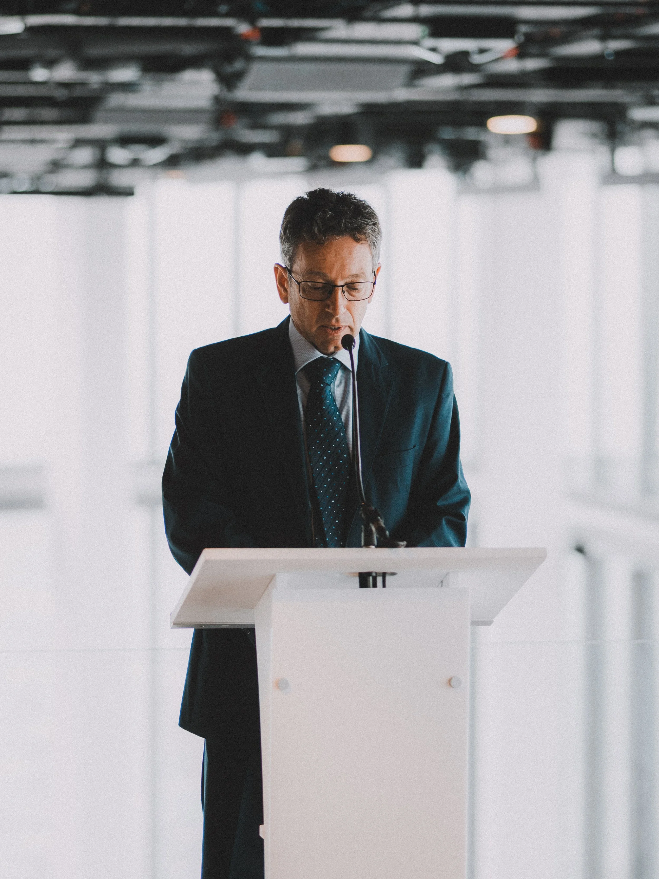A man in a dark suit and glasses standing at a white podium, reading or speaking into a microphone in a modern, bright indoor setting.