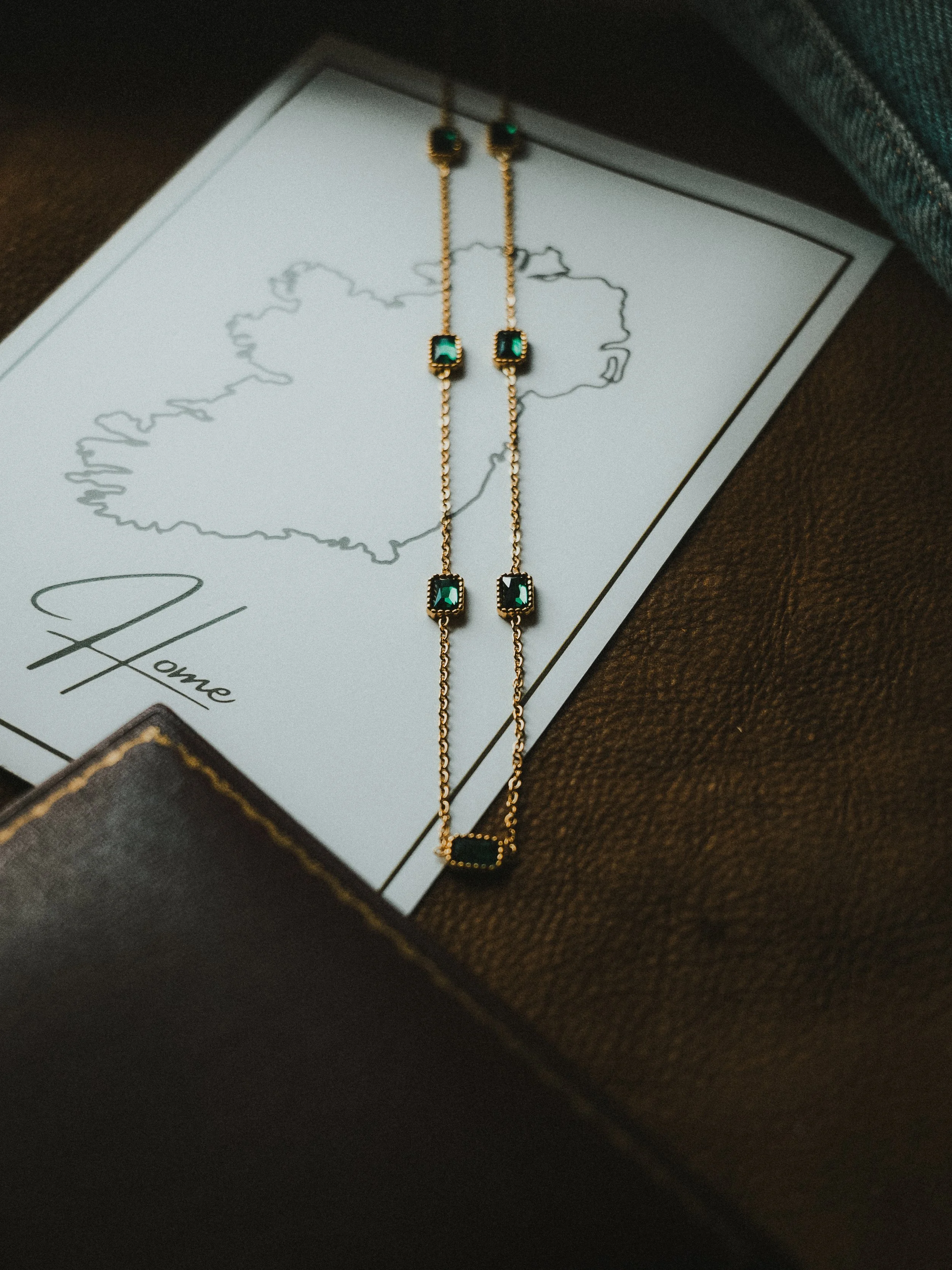 Gold chain necklace with small green stones, placed on a white card with a black outline of Italy, labeled 'Home', and partially resting on a dark brown leather surface.