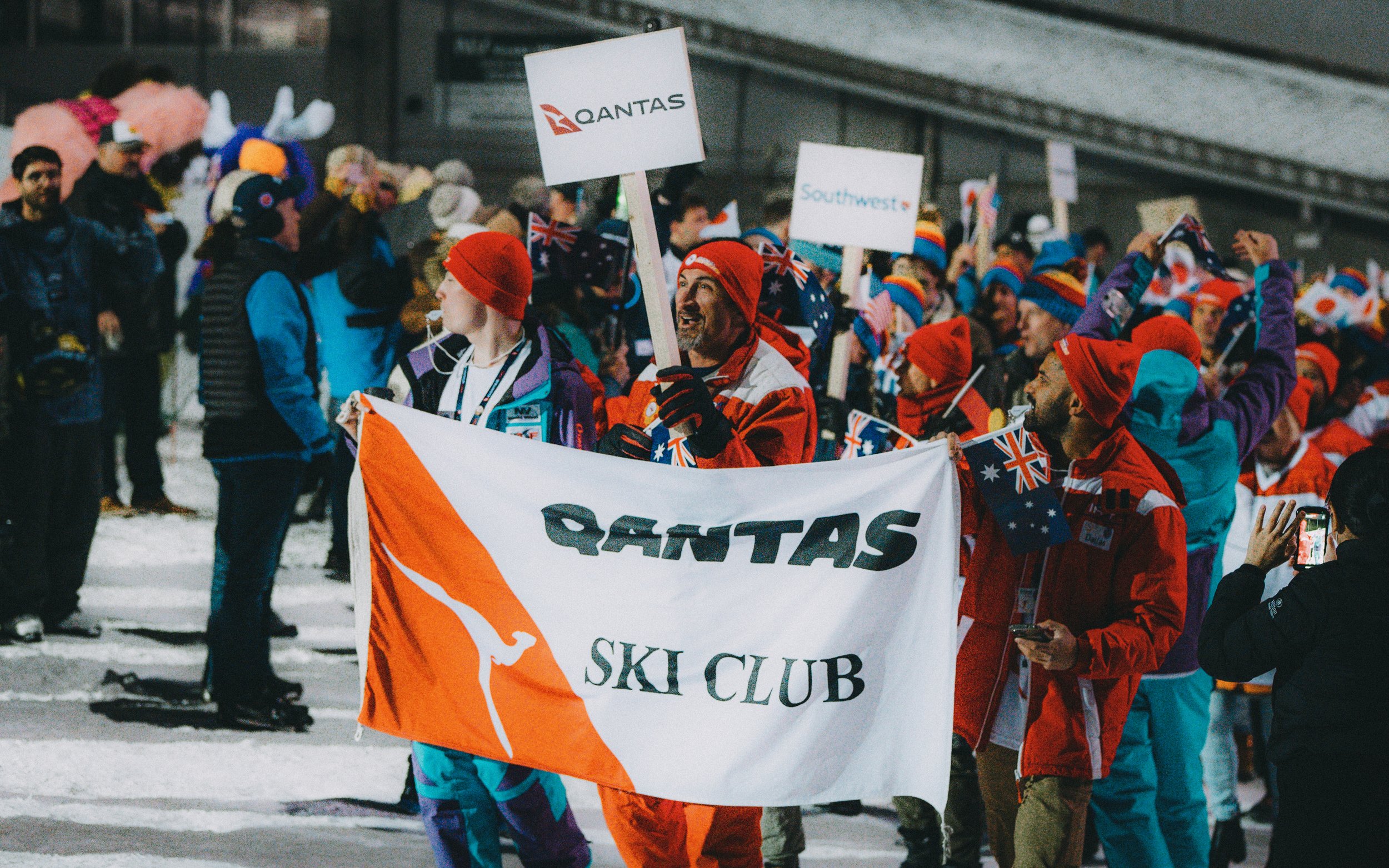 People participating in a winter sports event, holding a QANTAS Ski Club banner, wearing colorful winter clothing and hats, with some holding Australian flags.