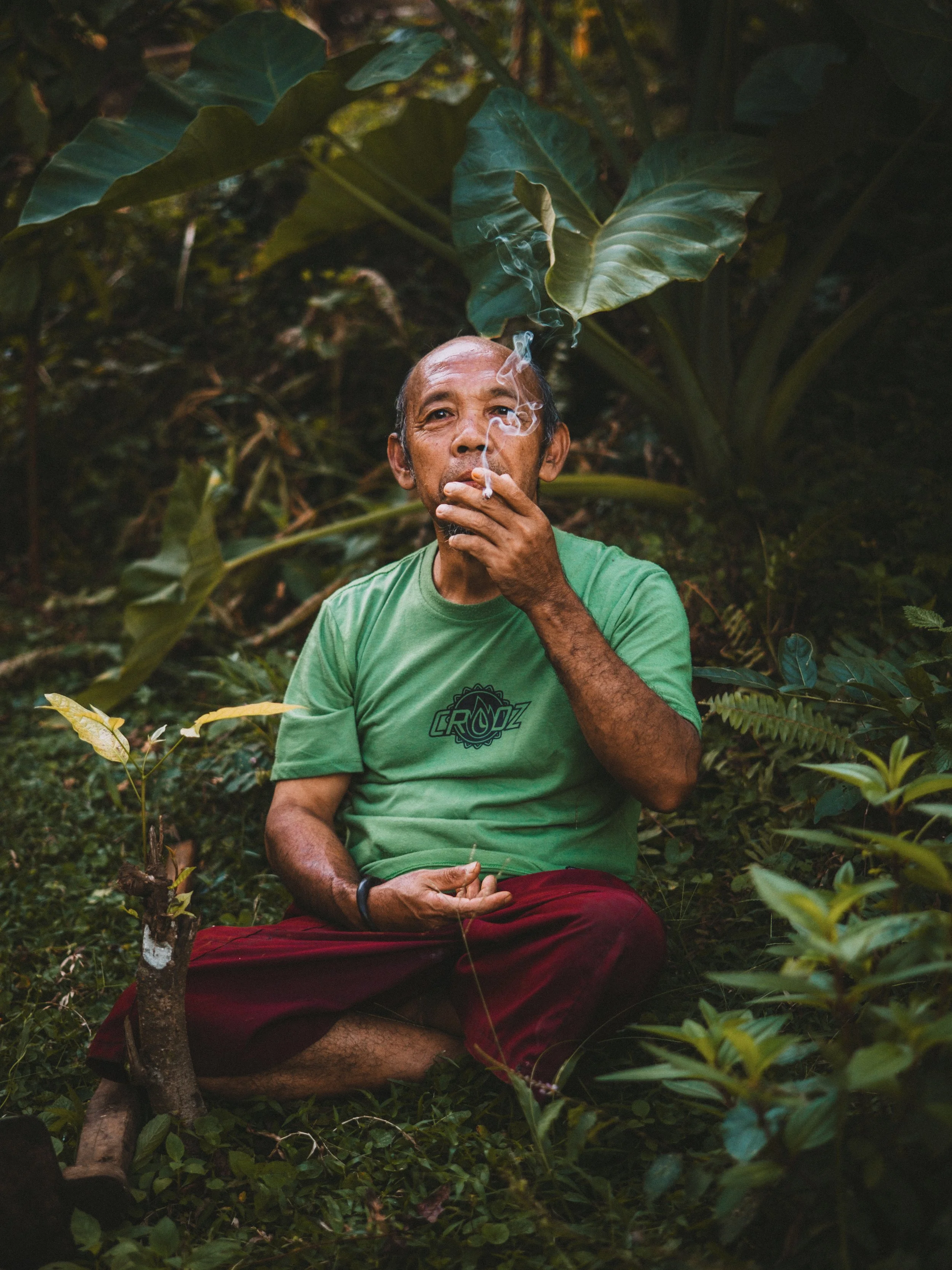 A man with dark skin, gray hair, and a beard sitting cross-legged in a dense, green jungle. He is wearing a green t-shirt with a logo and red pants, and is smoking a cigarette, with smoke rising from his mouth.