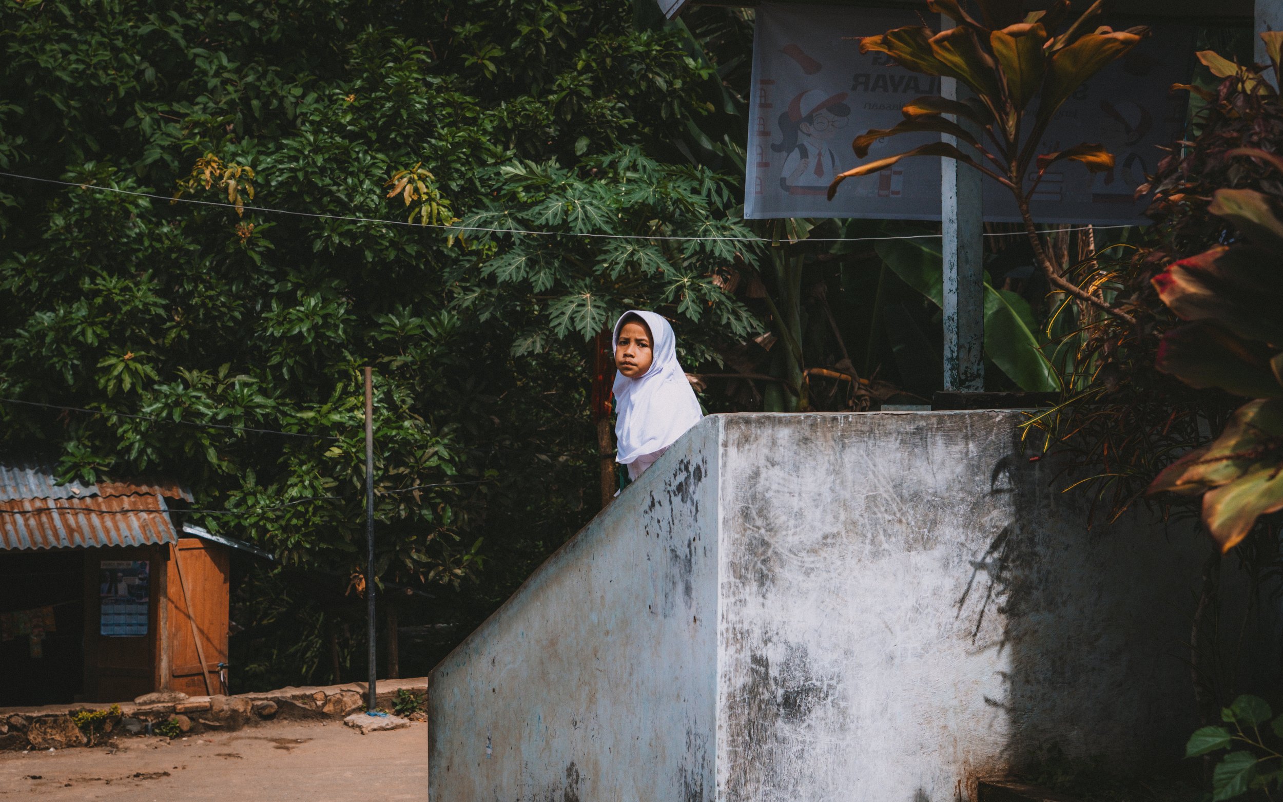 A young girl in a white hijab standing on a concrete platform, looking at the camera in a rural setting with dense green foliage and a small wooden structure behind her.