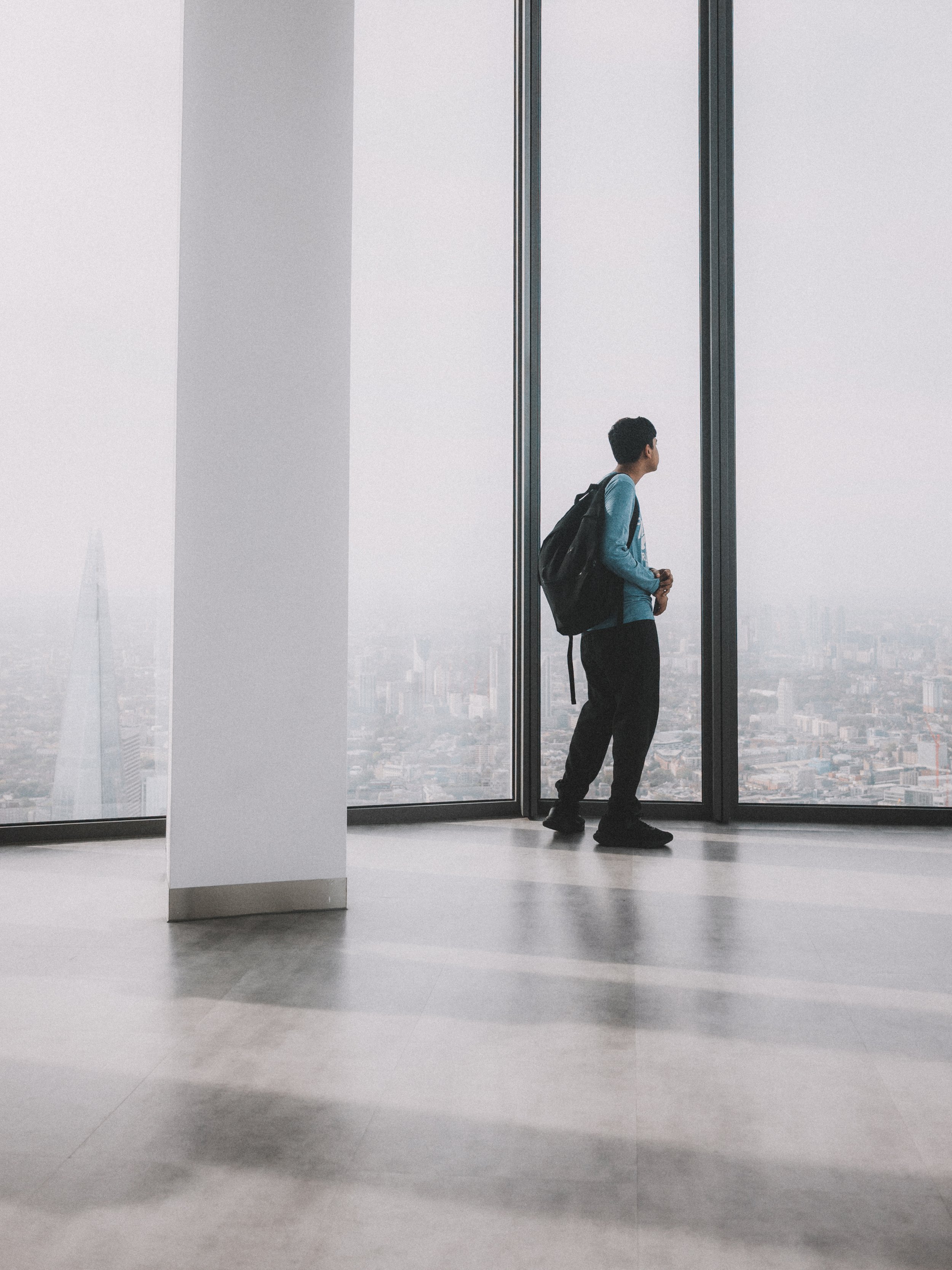 A young man with a backpack looking out the large windows of a high-rise building, overlooking a cityscape with tall buildings.