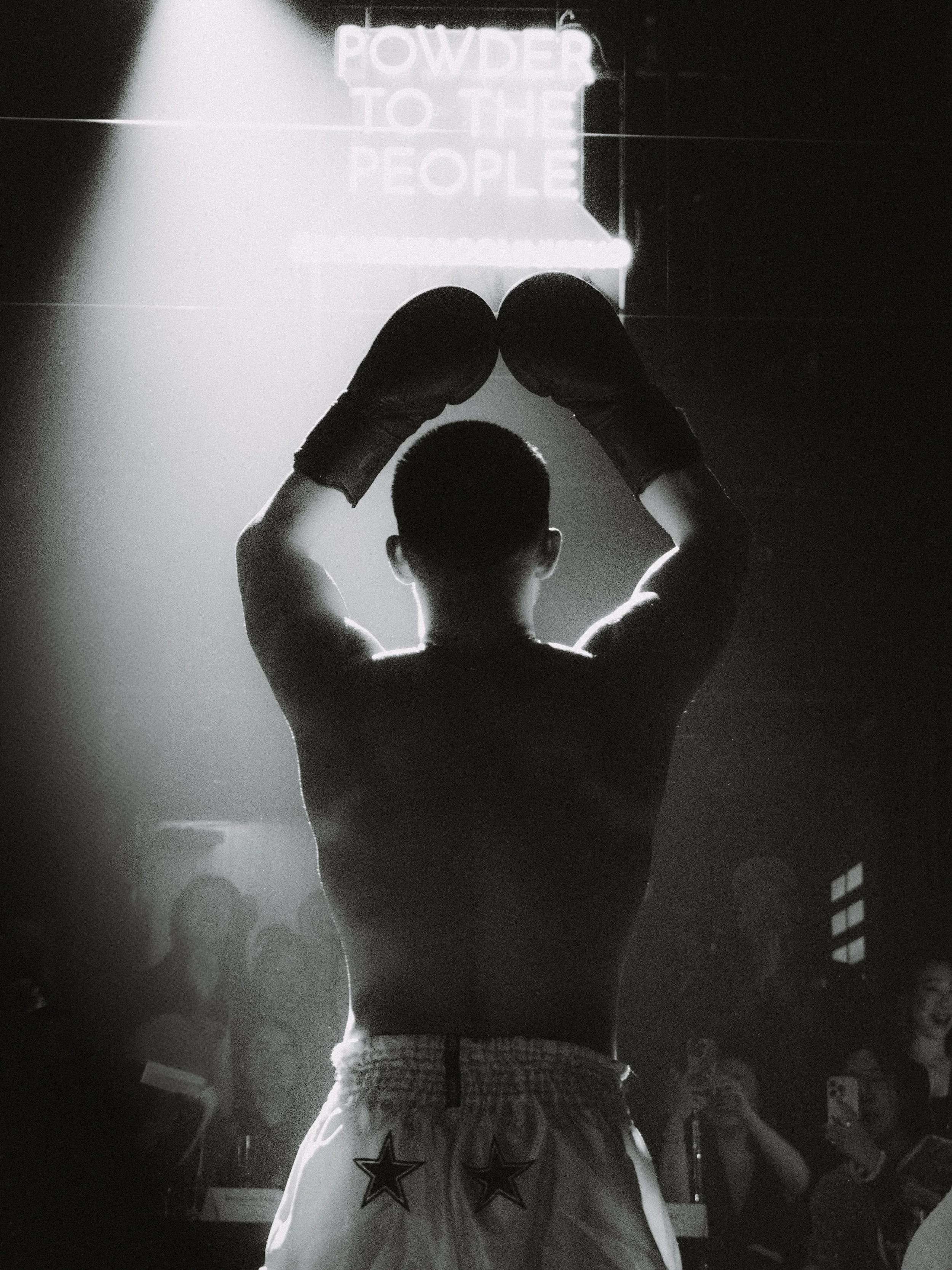 A boxer is standing with his back to the camera, forming a heart shape with his gloved hands above his head. Behind him is a bright neon sign that says 'POWDER TO THE PEOPLE.' An audience is watching and taking photos.