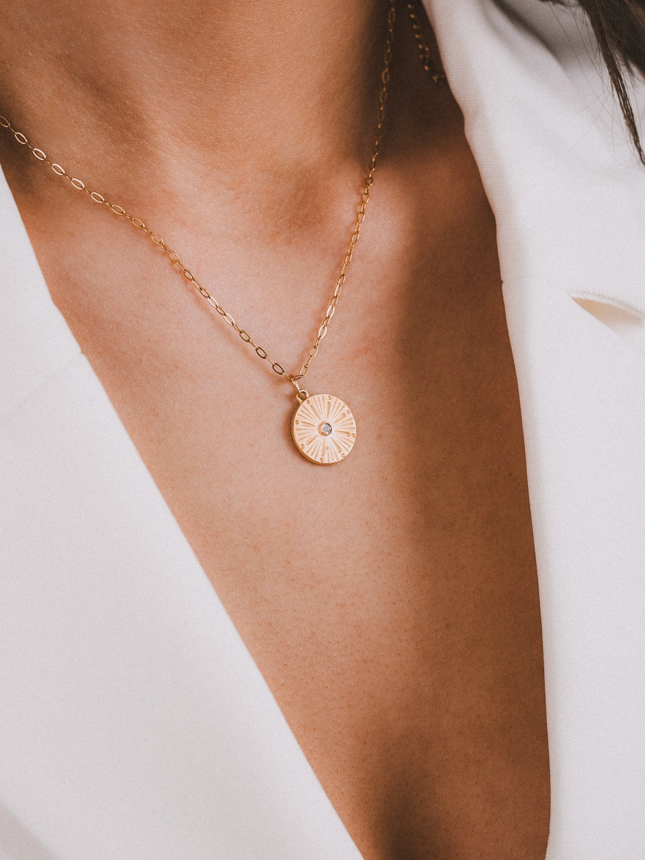 Close-up of a woman's neck and chest wearing a gold necklace with a round pendant featuring a small gemstone in the center