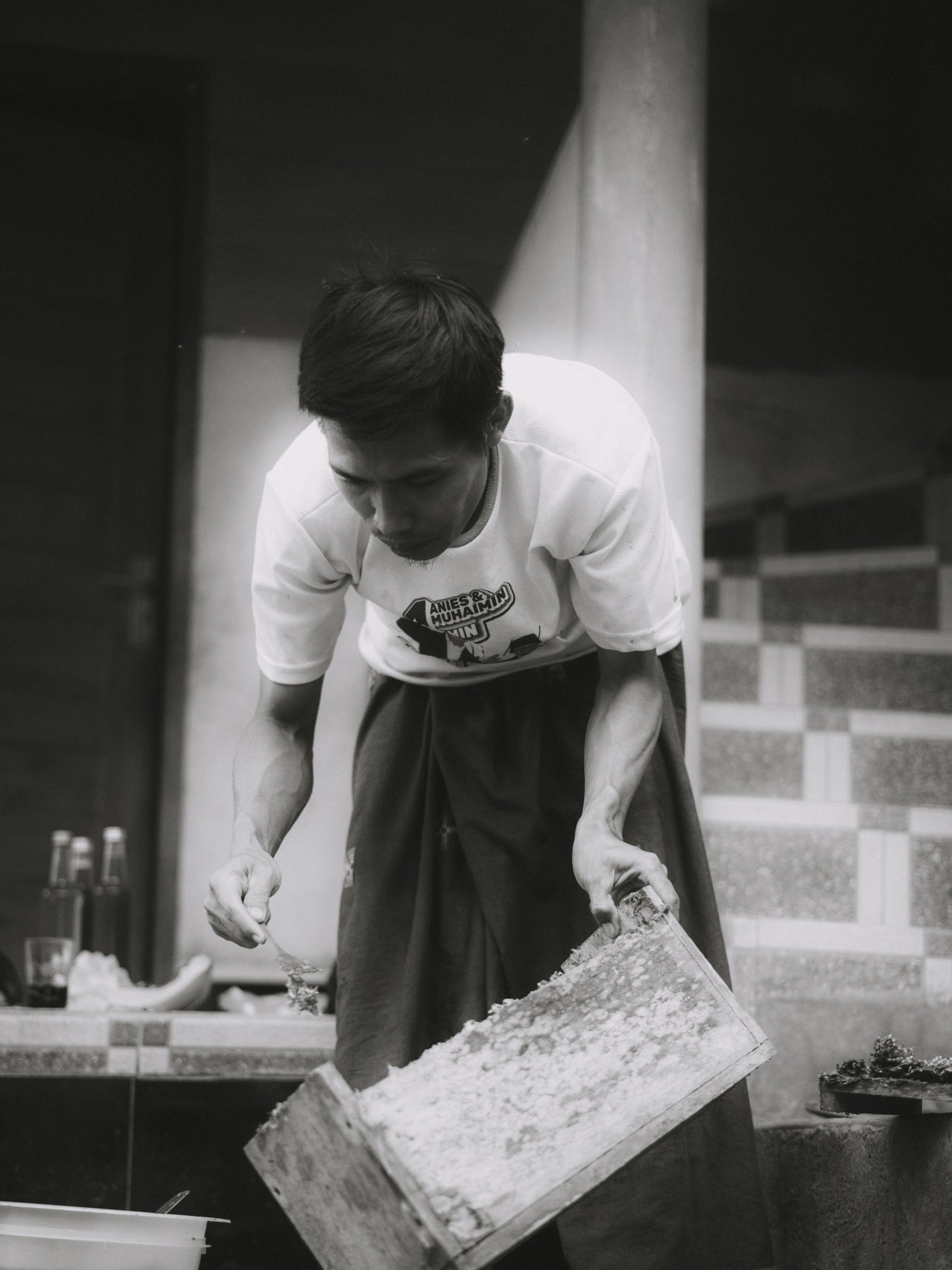 A man wearing a white t-shirt and dark pants is pouring rice from a wooden container into a bowl indoors.
