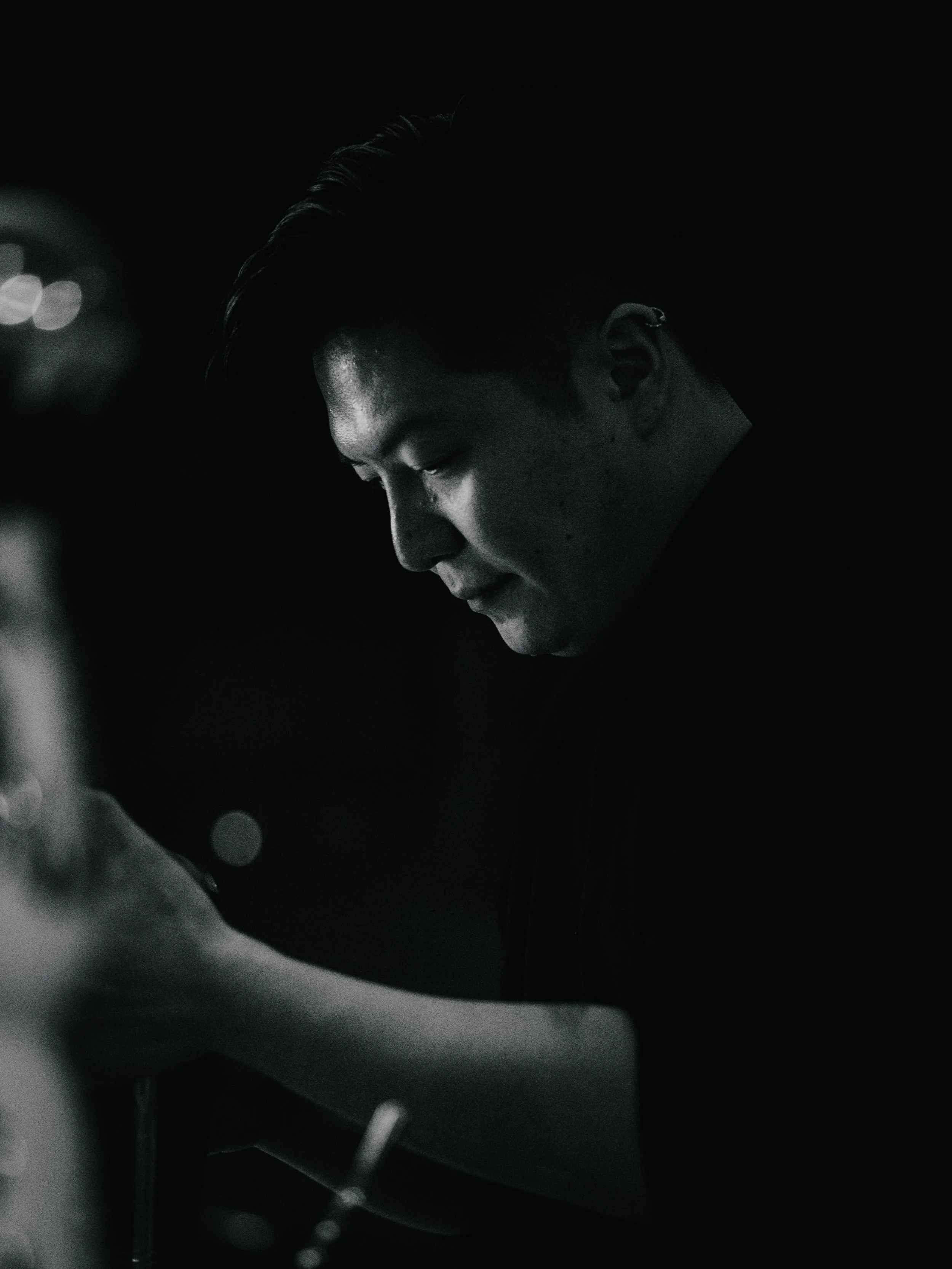 A black and white photo of a young man with short, styled hair, looking downwards with a pensive expression, in a dark environment.