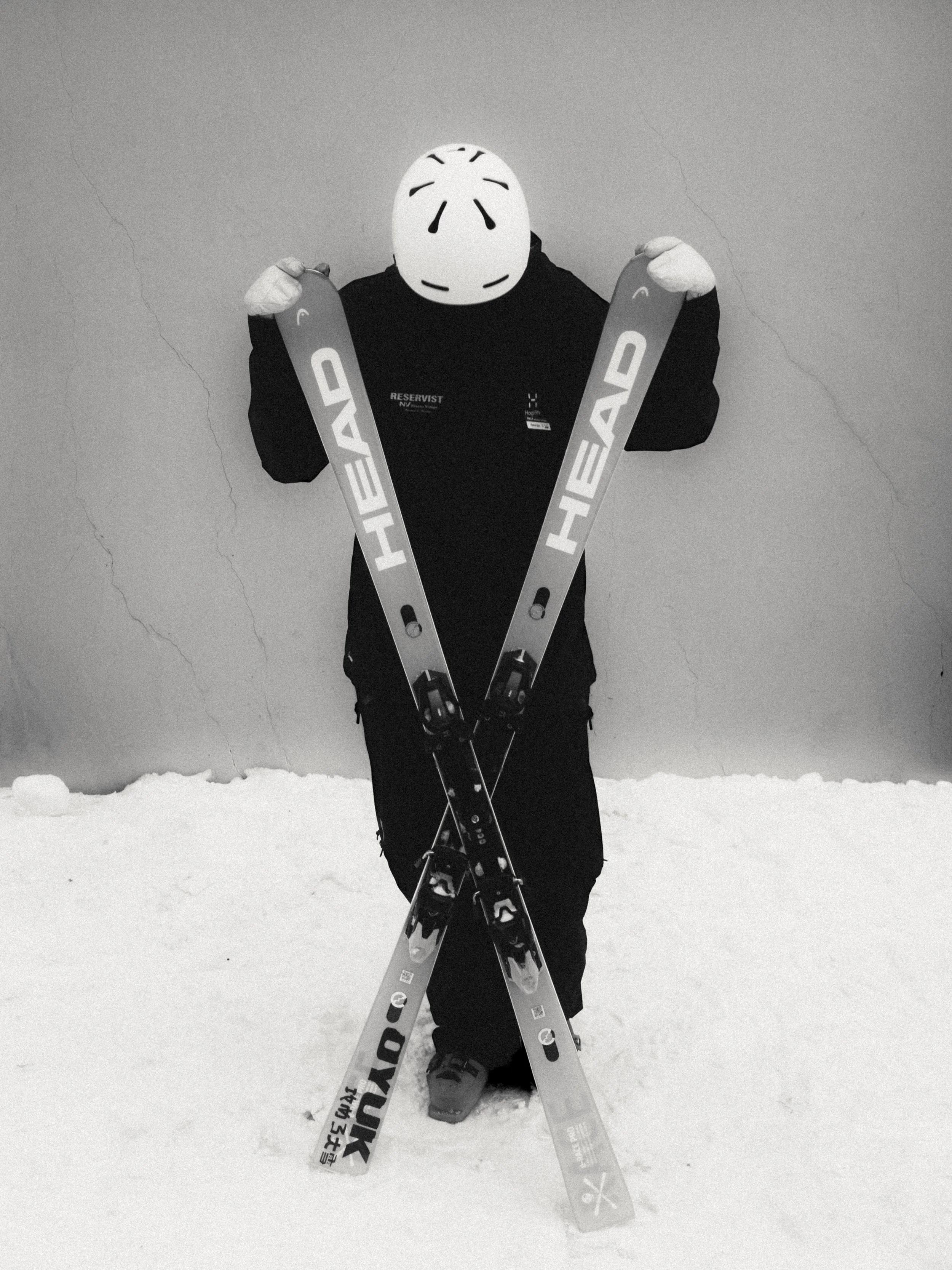 Person wearing a ski mask holding crossed skis on a snowy surface indoors.
