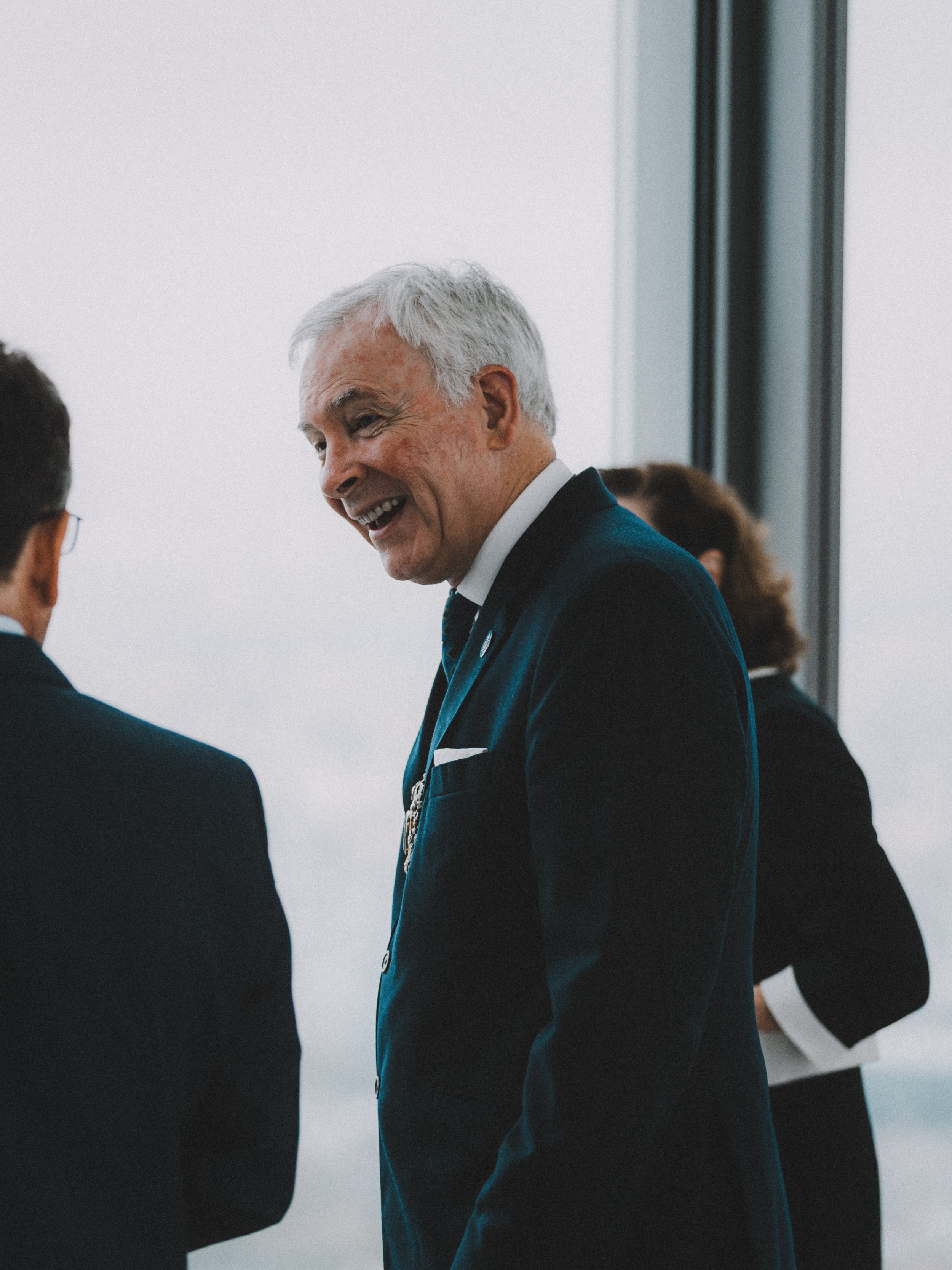 An older man with gray hair smiling and talking to someone, dressed in a dark suit, indoors near large windows.