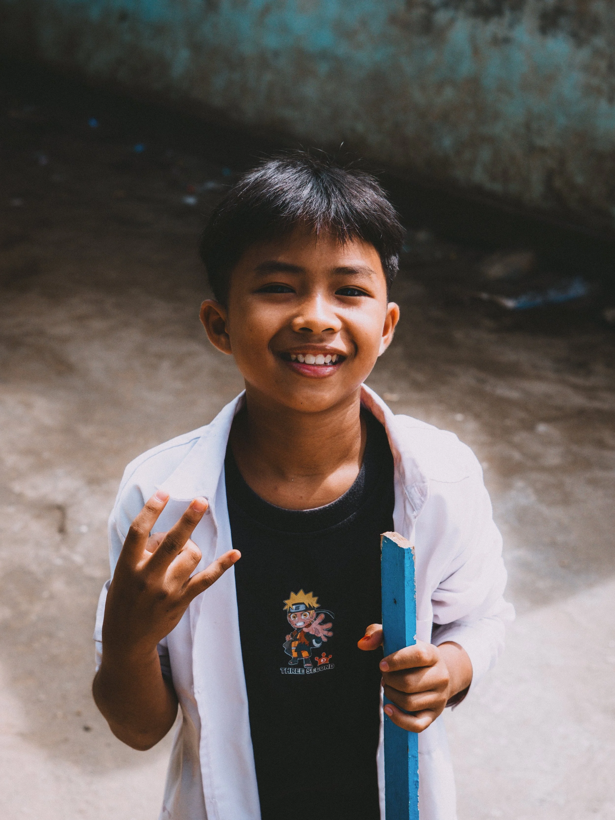A smiling boy holding a blue chalkboard, making a gesture with his hand, wearing a black Naruto T-shirt and an open white shirt, standing outdoors.