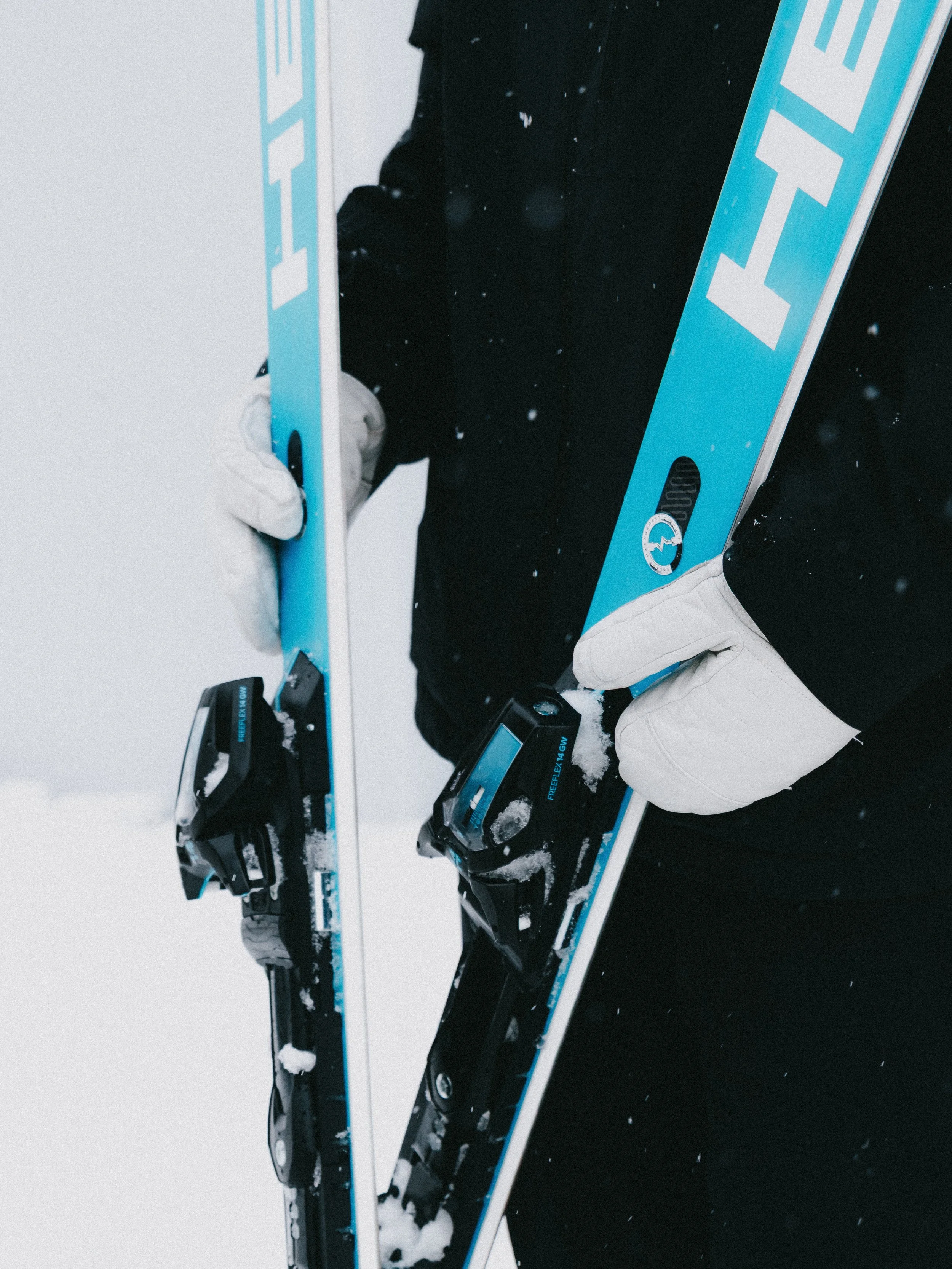 Person holding skis and ski bindings in snowy environment, wearing gloves and dark winter clothing.