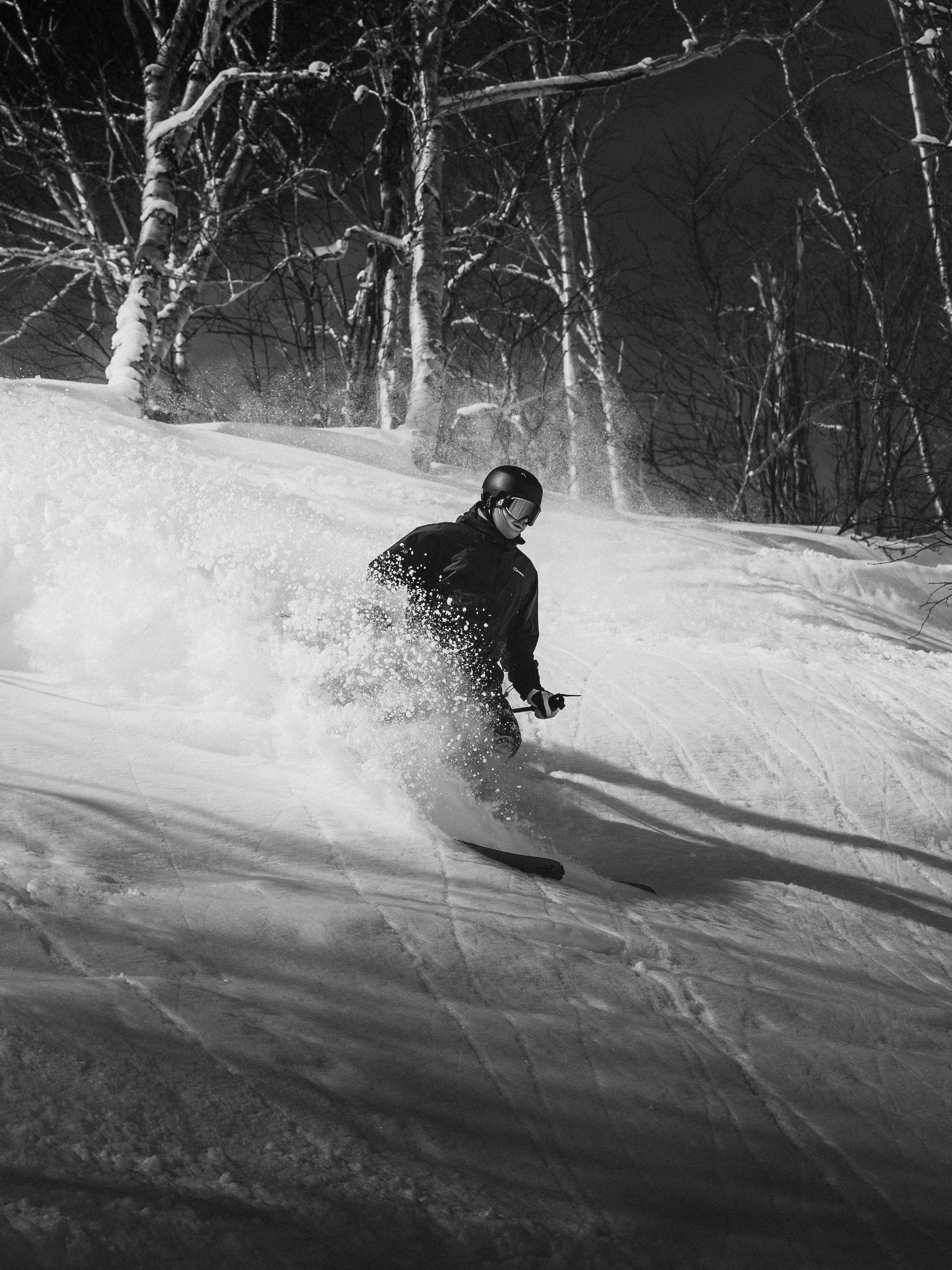 A person skiing downhill at night on a snow-covered slope, wearing a helmet and goggles, with bare trees in the background.