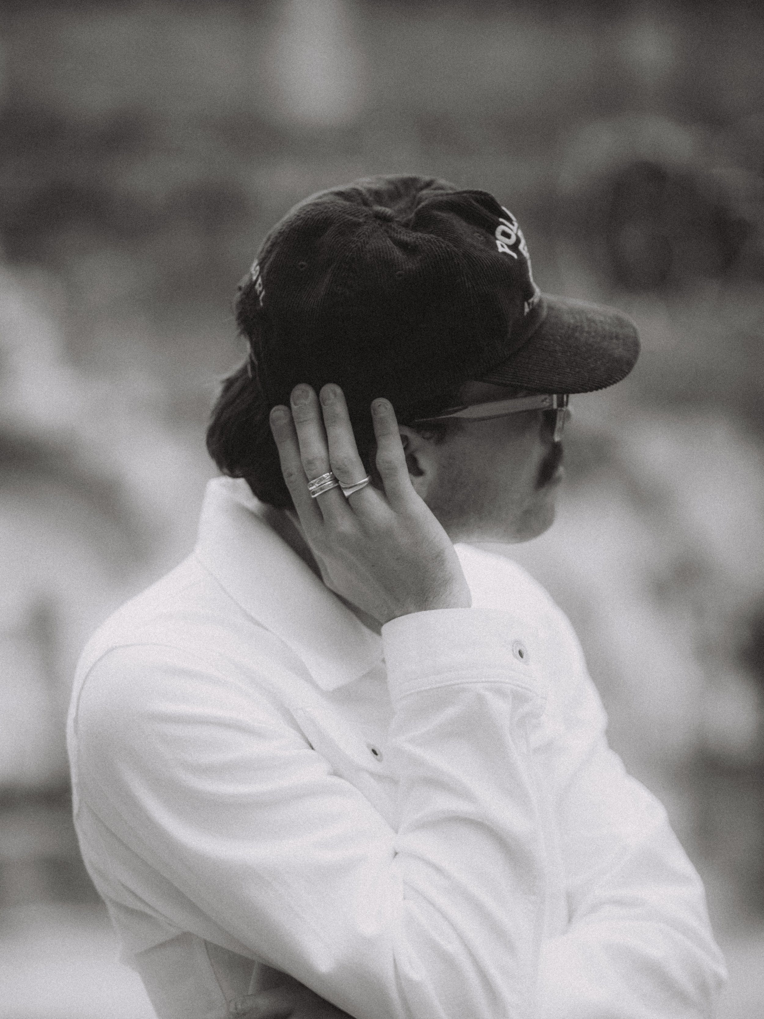 Side view of a person wearing sunglasses, a white jacket, a dark cap, and rings on their fingers, holding the back of their head with one hand in black and white image.