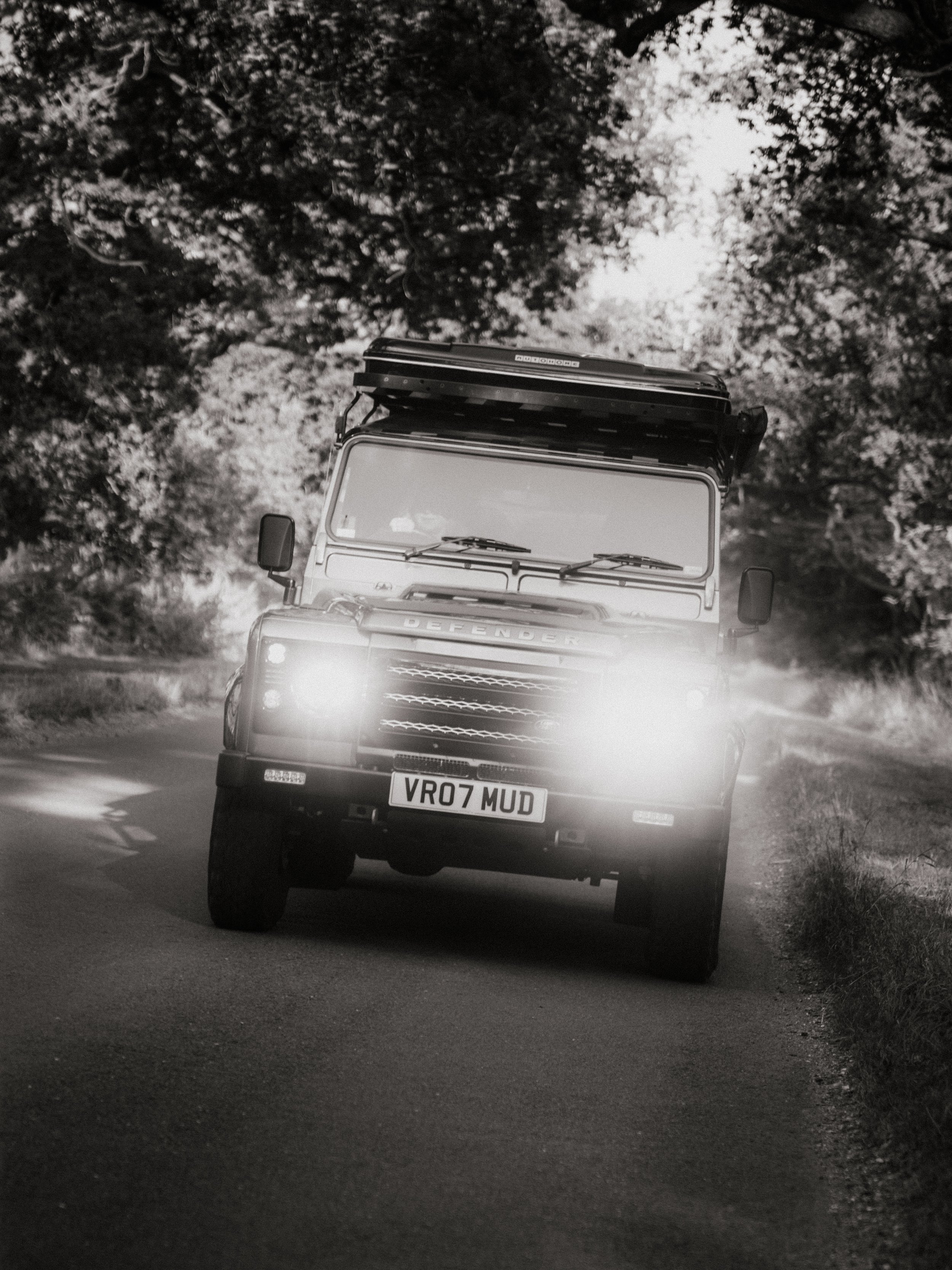 A black-and-white photo of a Land Rover Defender driving on a curved rural road surrounded by trees, with bright headlights shining forward.