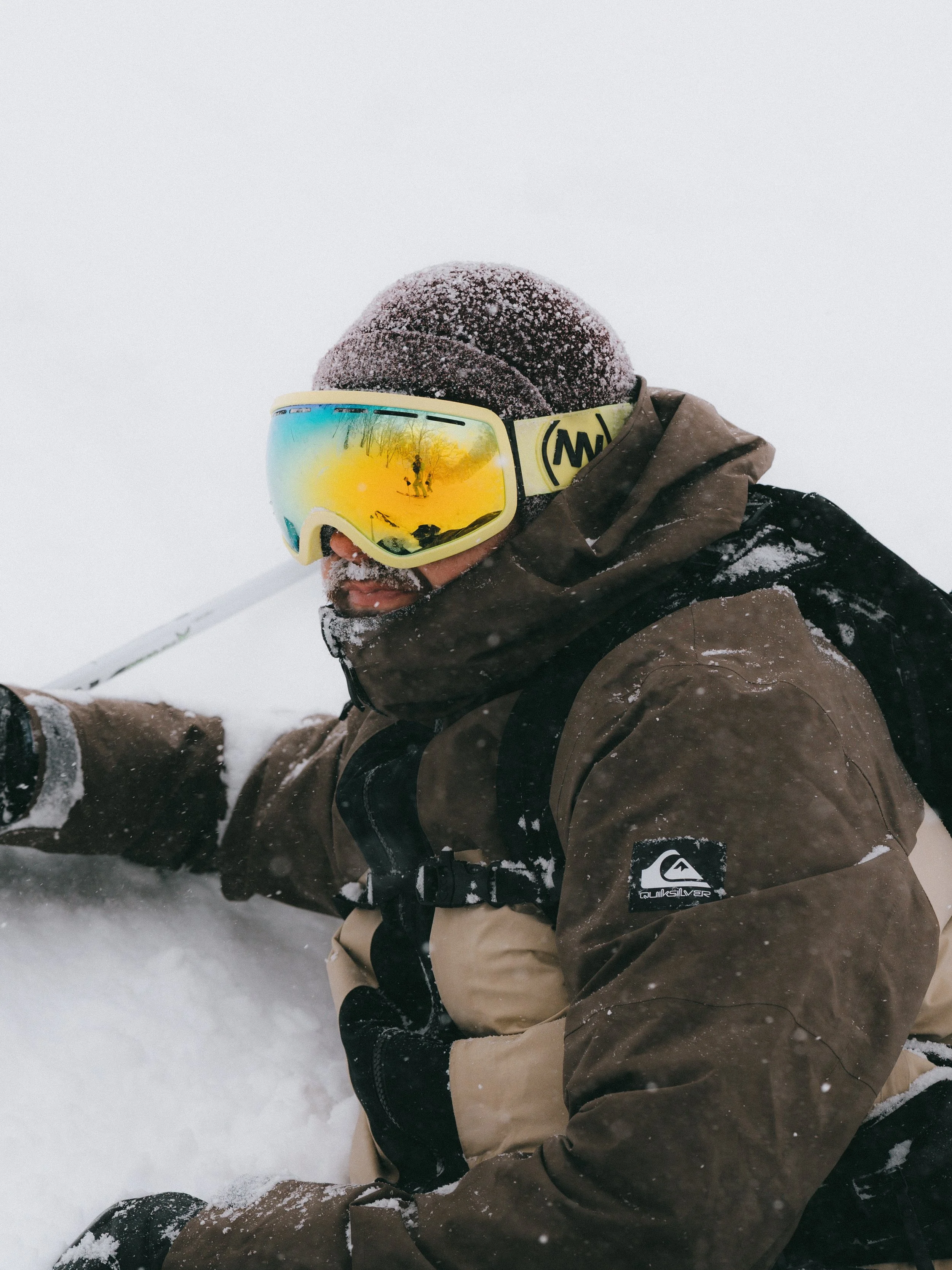 Person wearing a brown winter jacket, yellow goggles with a reflection of a snowy landscape, a brown hat, and gloves, sitting in the snow during a snowstorm.