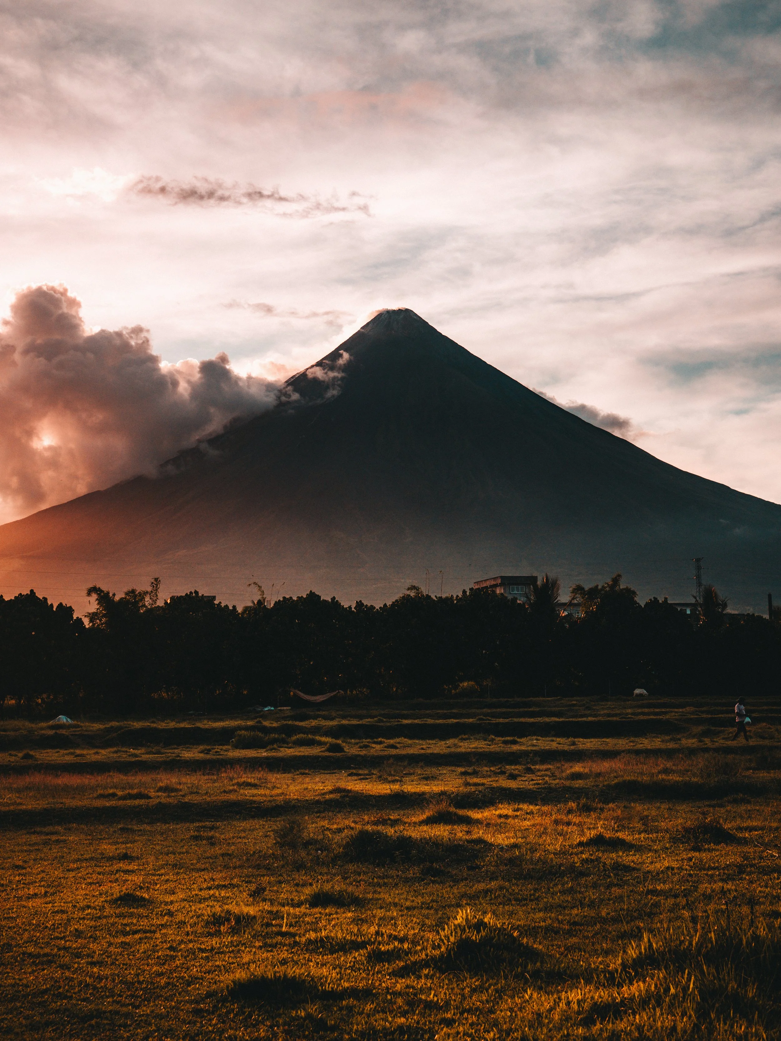 A mountainous volcano during sunset with clouds in the sky and a person running on grassy field in the foreground.