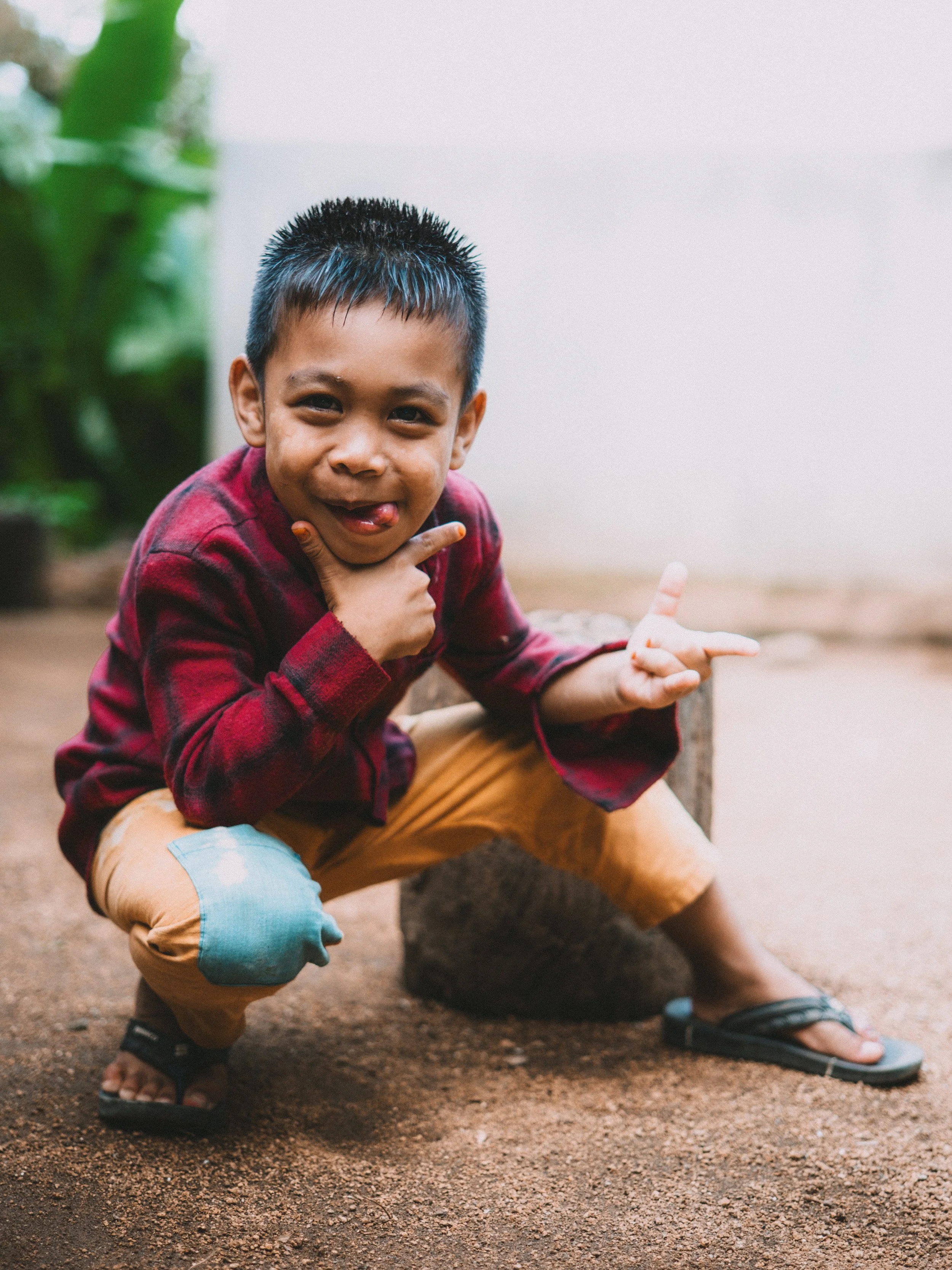 A young boy crouching outdoors, making a playful gesture with his hand, smiling with his tongue out, and wearing a red plaid shirt, tan pants with a patch, and black flip-flops.