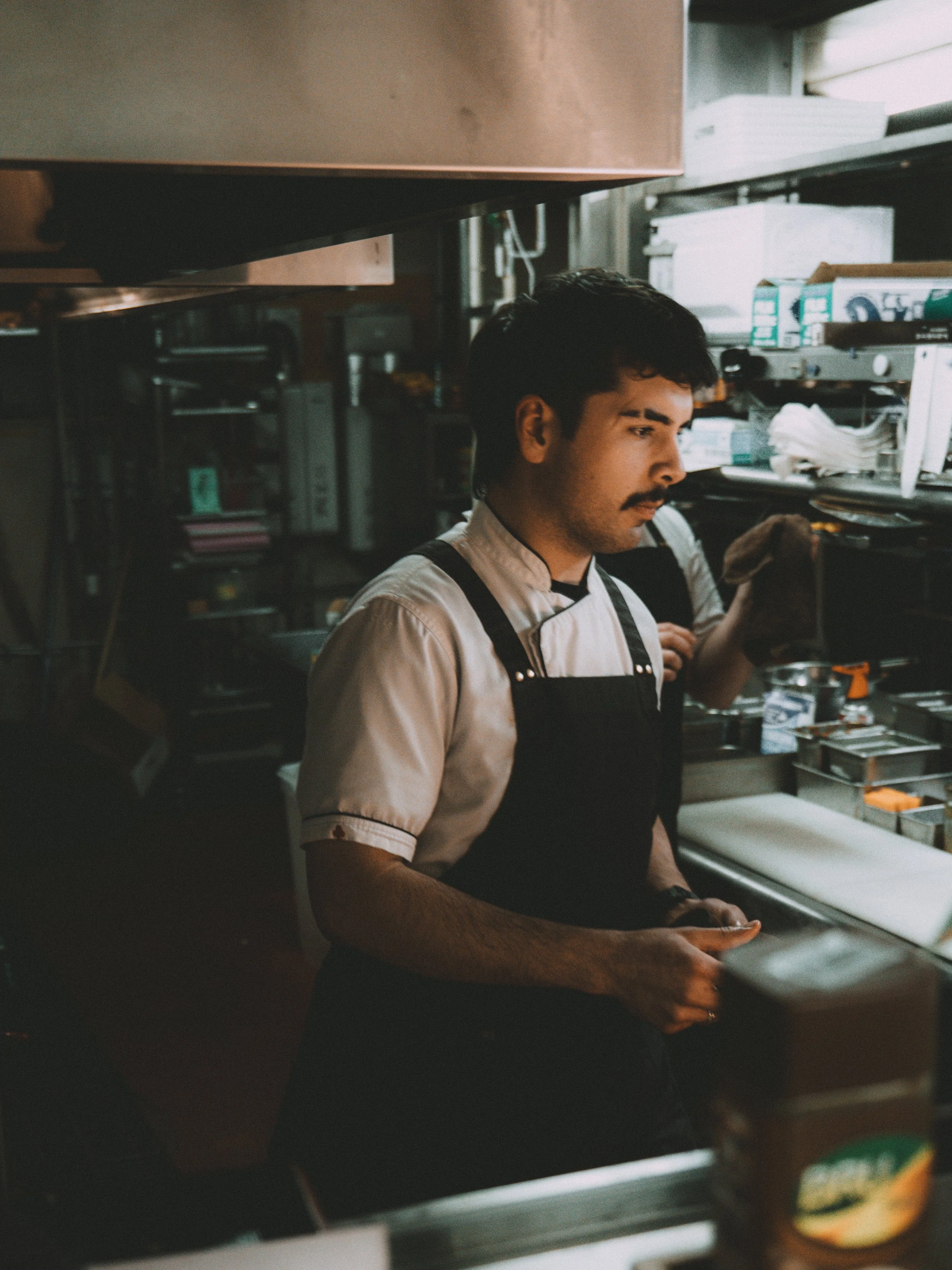 Male chef in white shirt and black apron working in a busy kitchen.