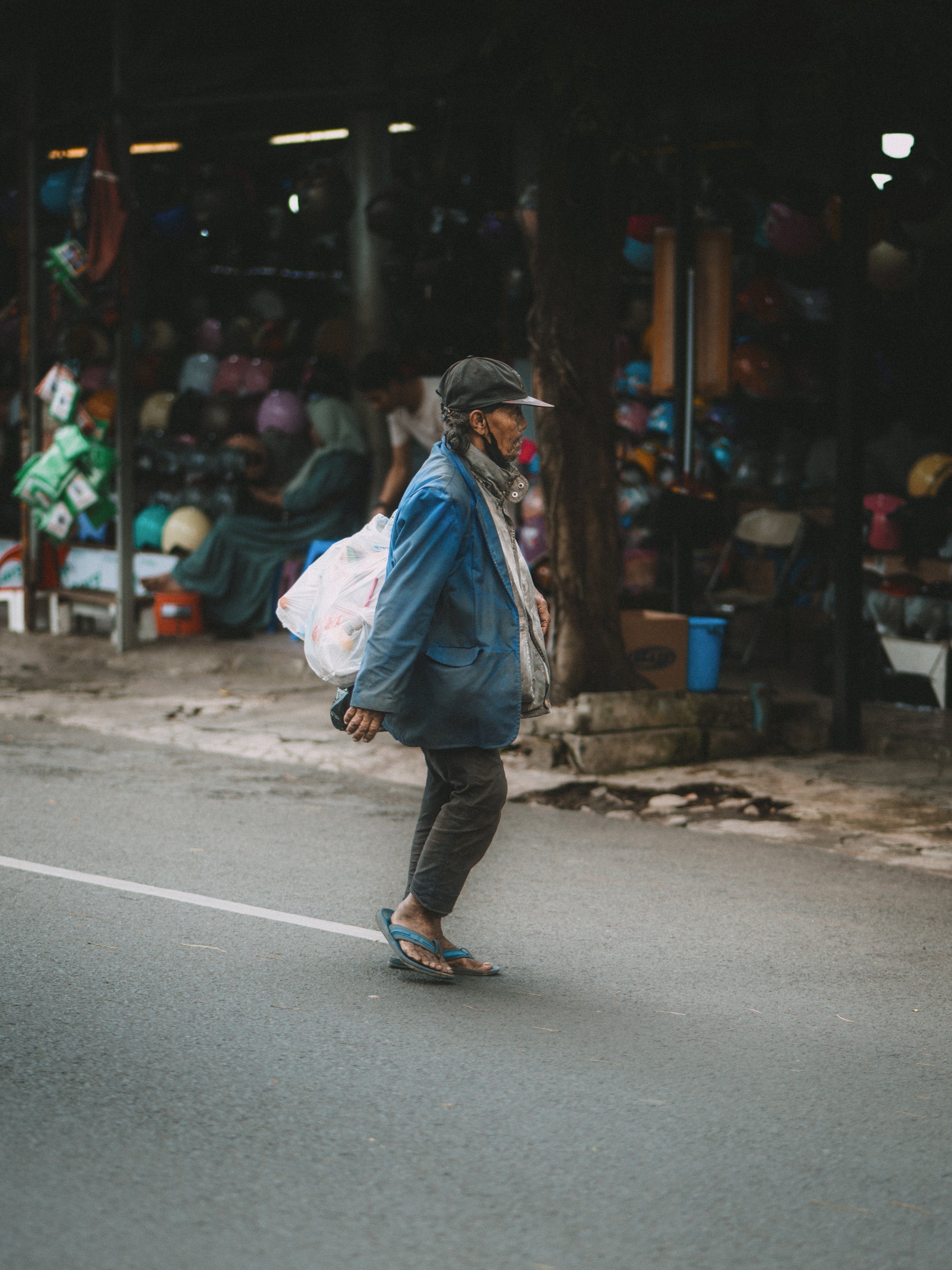 Older man walking on the street with a plastic bag over his shoulder, wearing a blue jacket, black cap, and flip-flops.