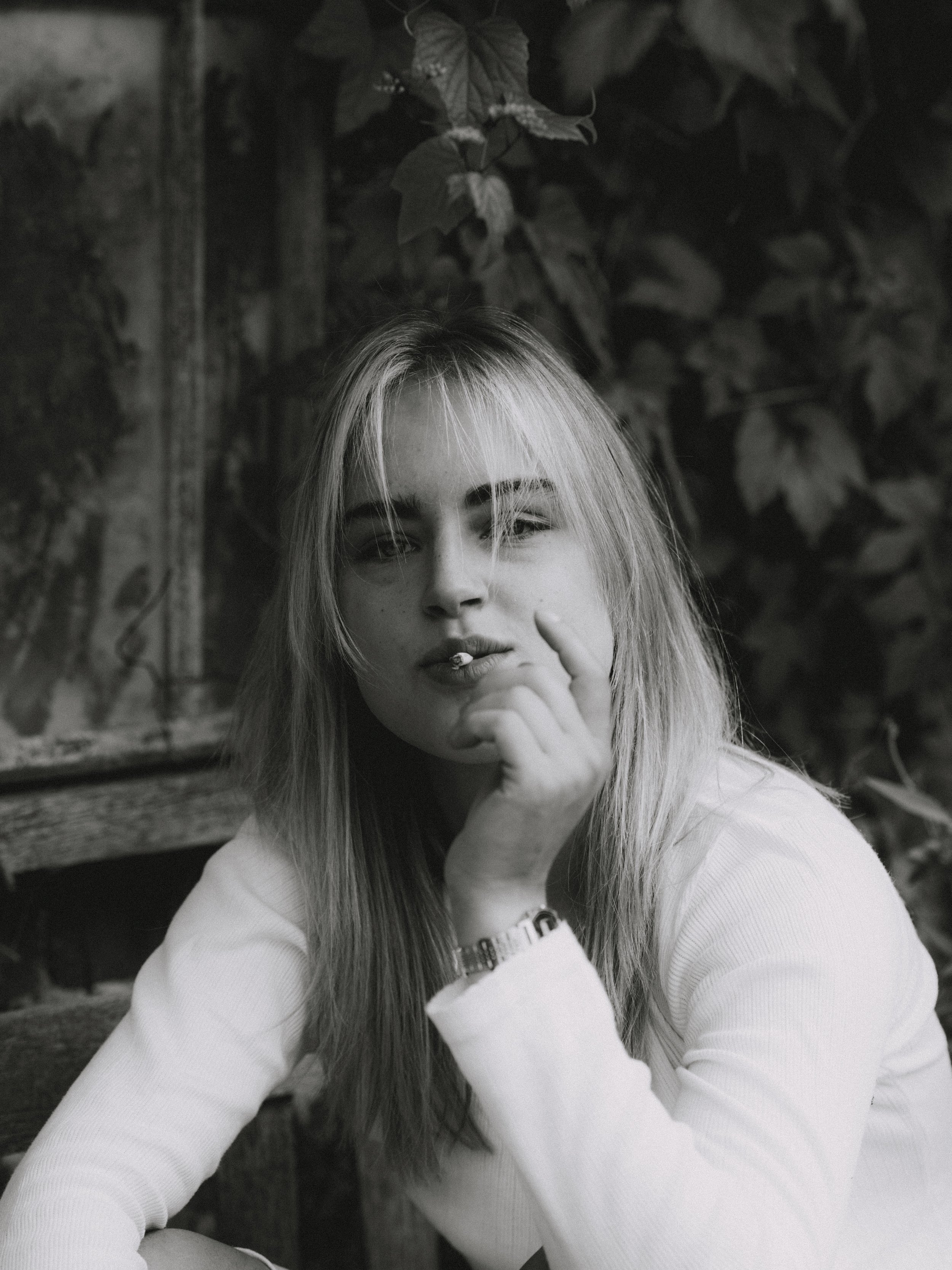 Black and white photo of a young woman with long blonde hair, wearing a white top, resting her chin on her hand, with a thoughtful expression, outdoors with foliage in the background.