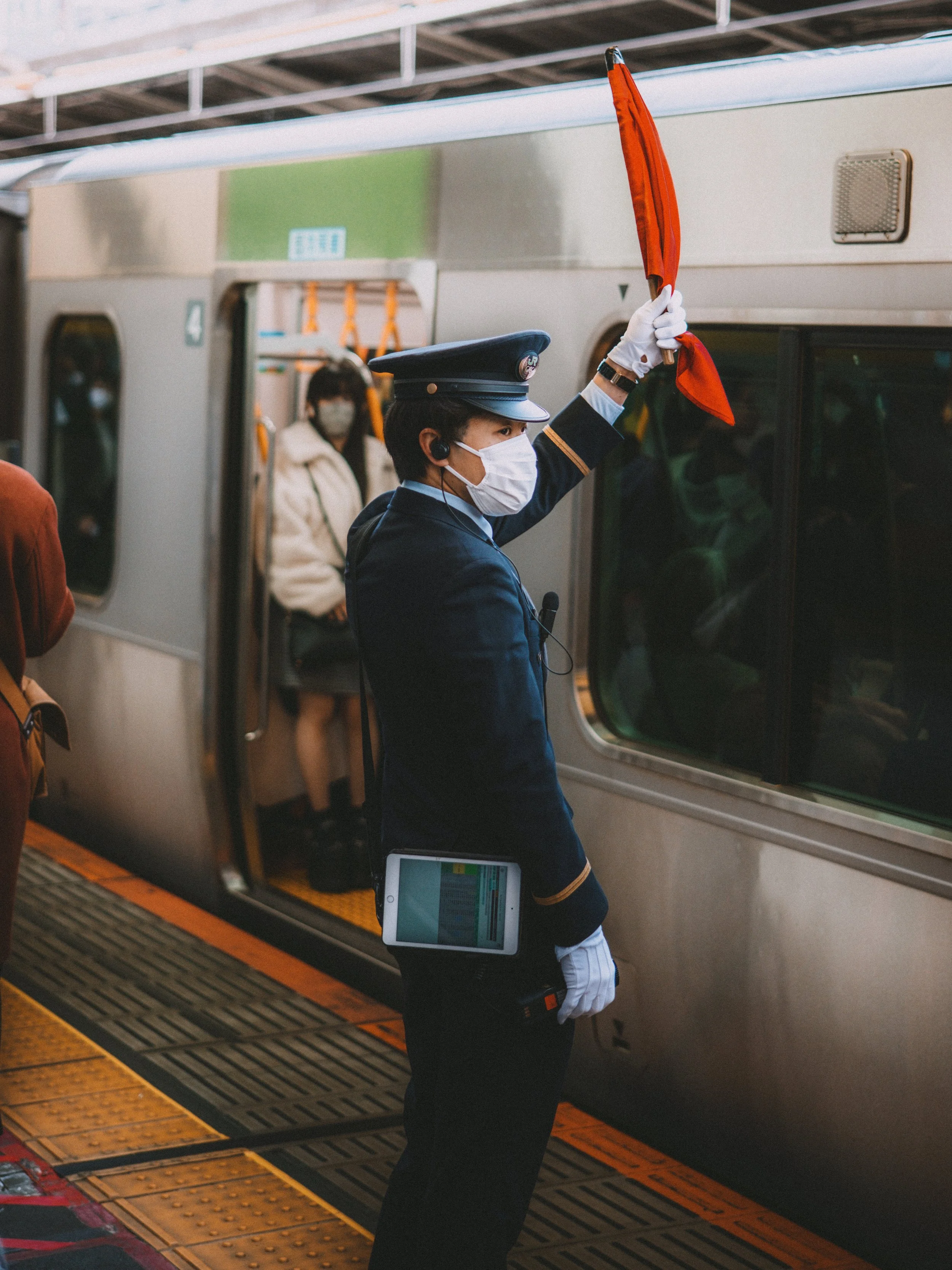 Train conductor in uniform and mask holding a red flag on a subway platform with passengers boarding a train.